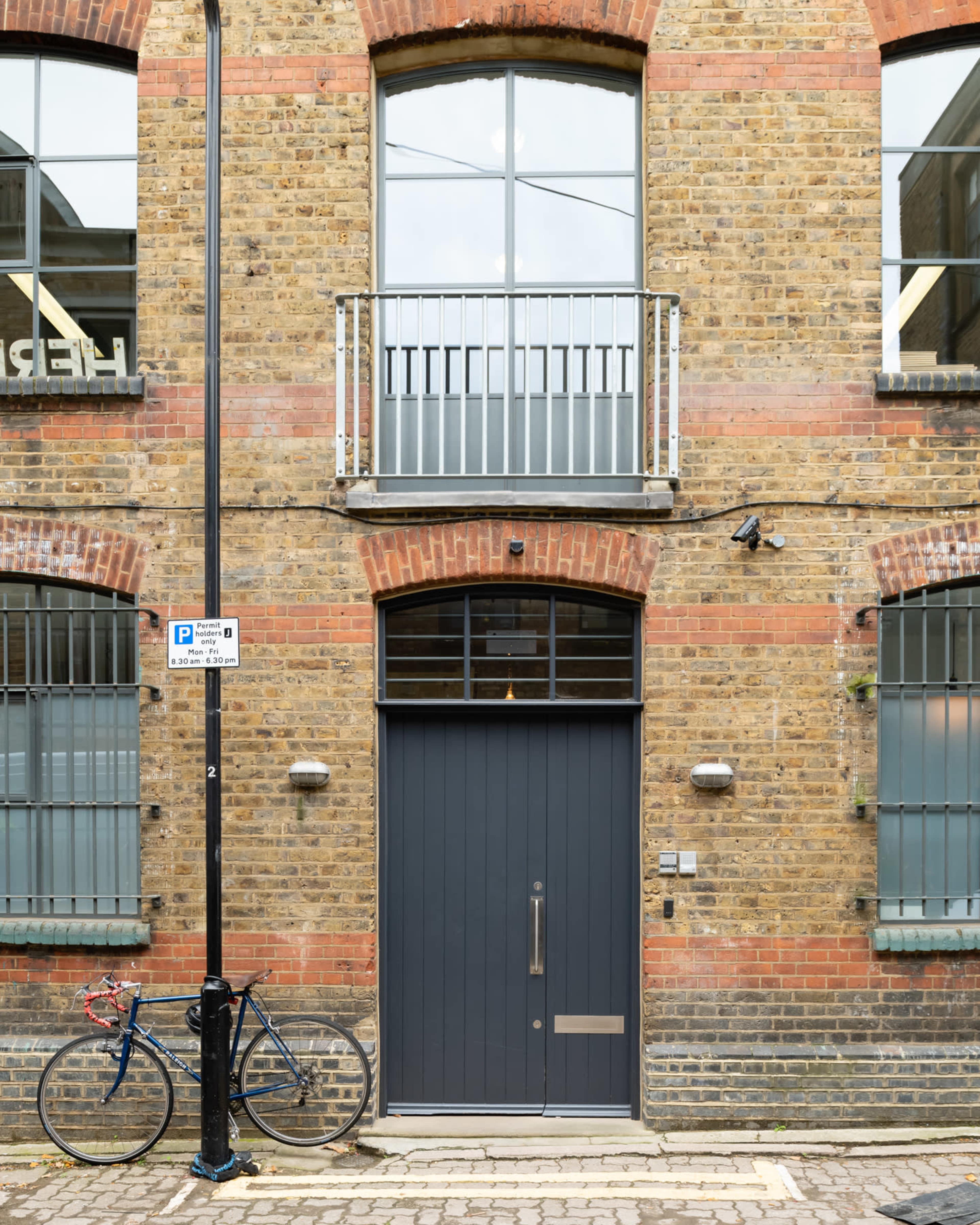 The entrance of a brick building featuring large windows above a dark door, with a bicycle parked beside a streetlight.