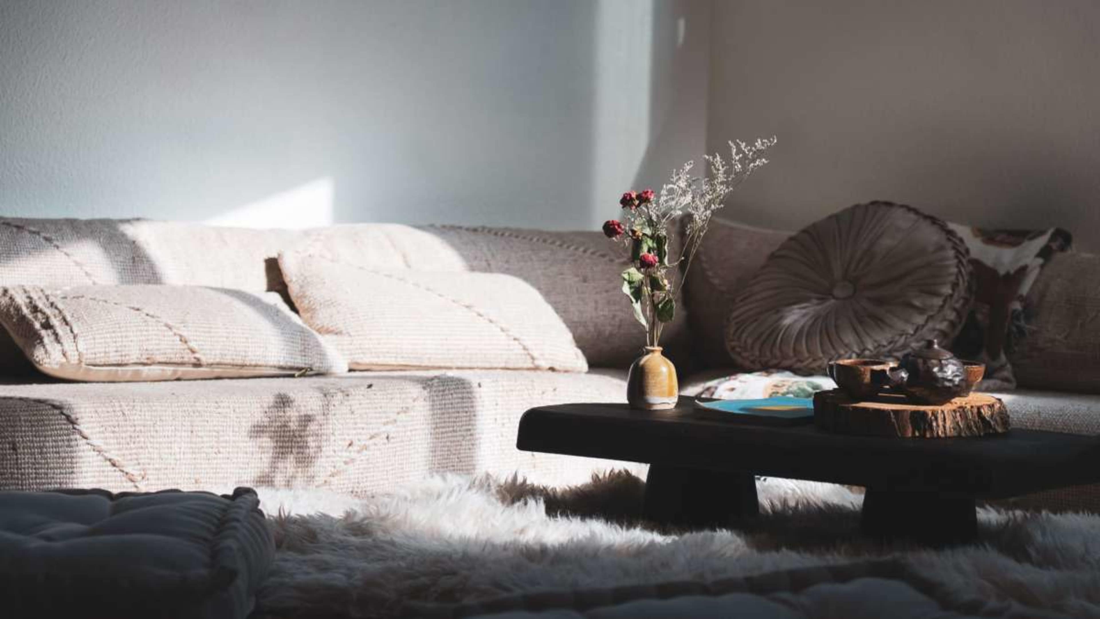A light-filled living room features a plush sofa with decorative pillows and a coffee table made of wood, adorned with a vase of dried flowers and a small decorative item.