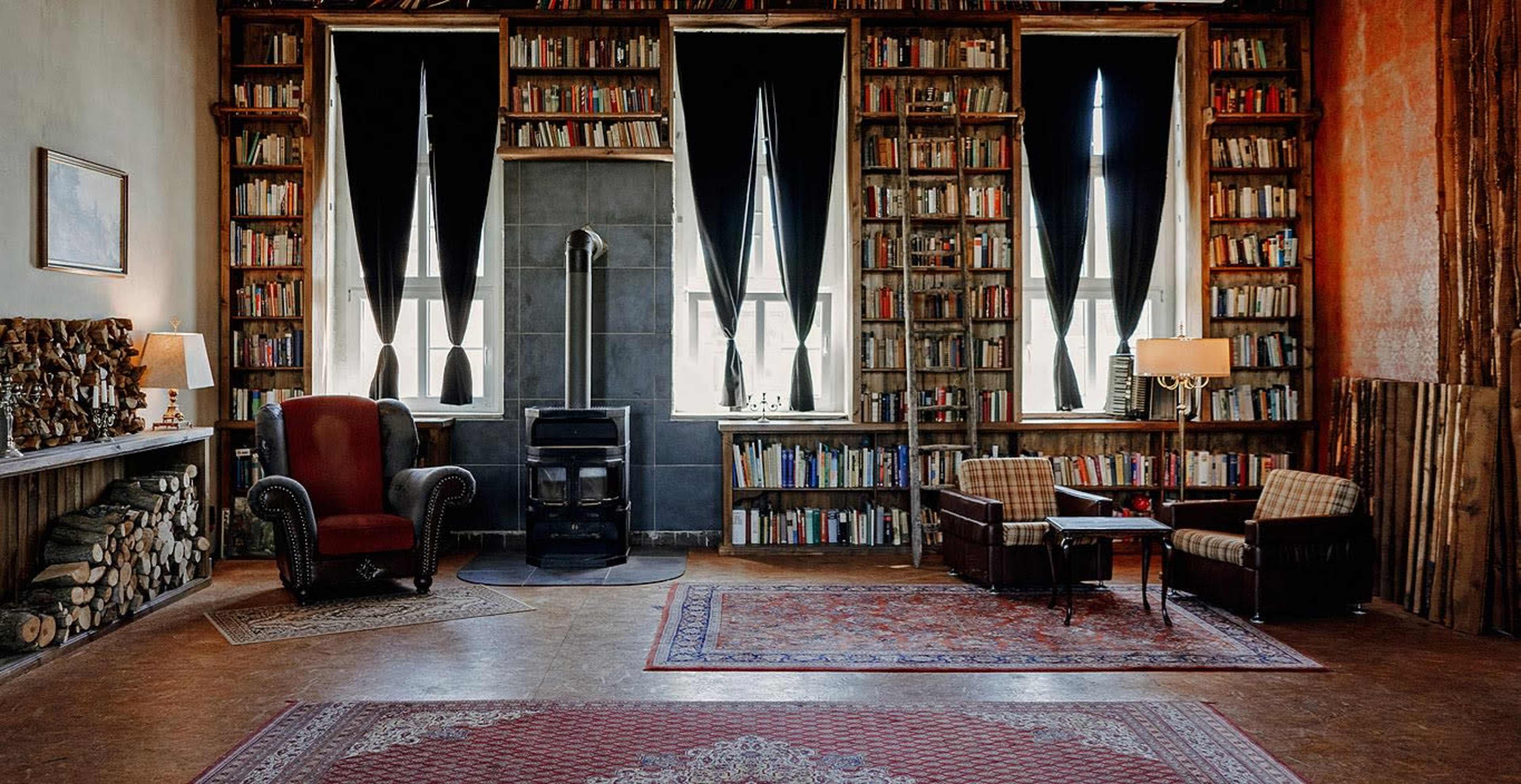 The image shows a cozy library with tall bookshelves filled with books, two armchairs, a small table, and a fireplace with a stack of logs beside it.