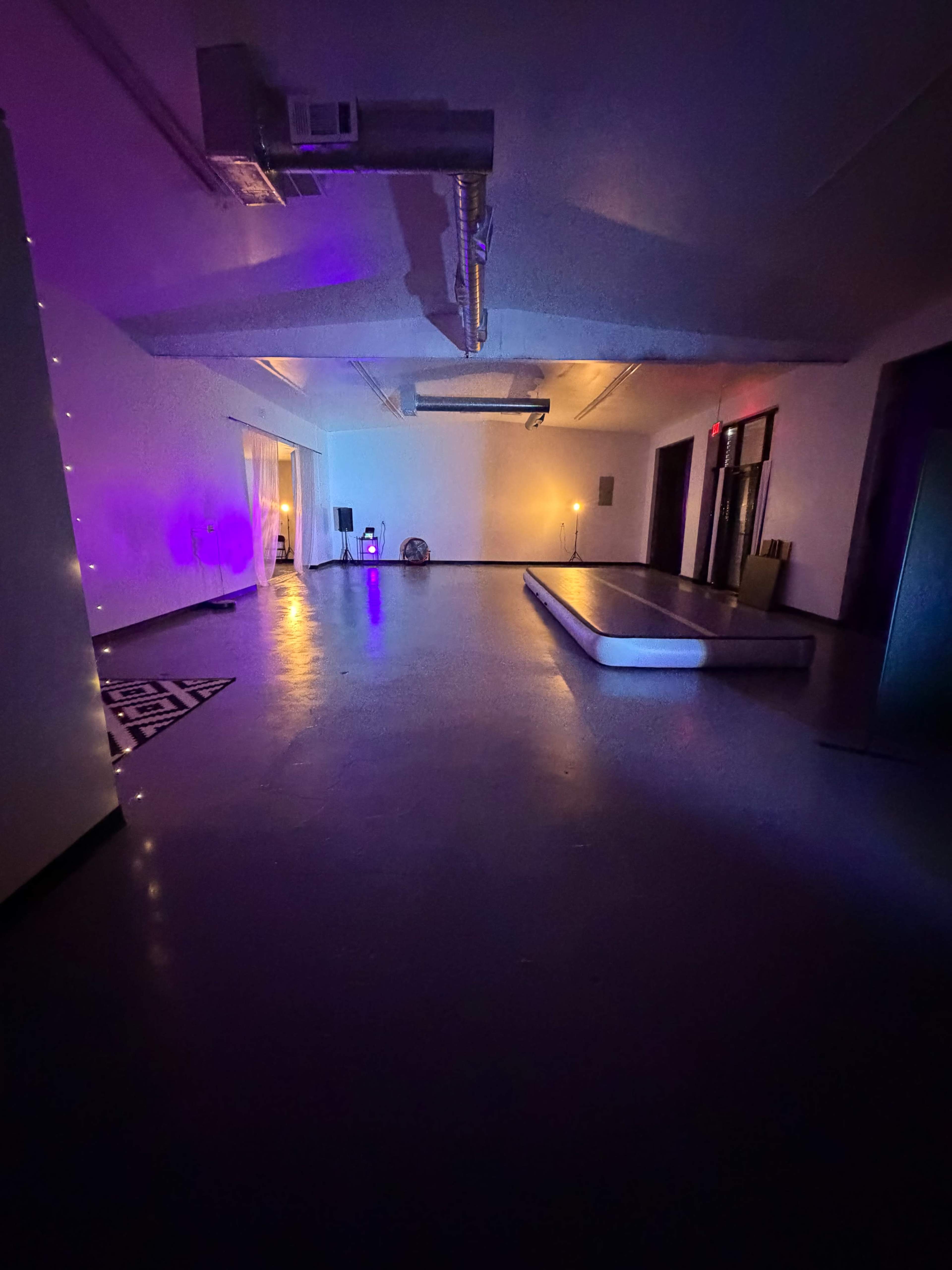 The image shows a spacious, dimly lit room with purple and orange lighting, featuring a partially visible platform and minimal furniture against the walls.