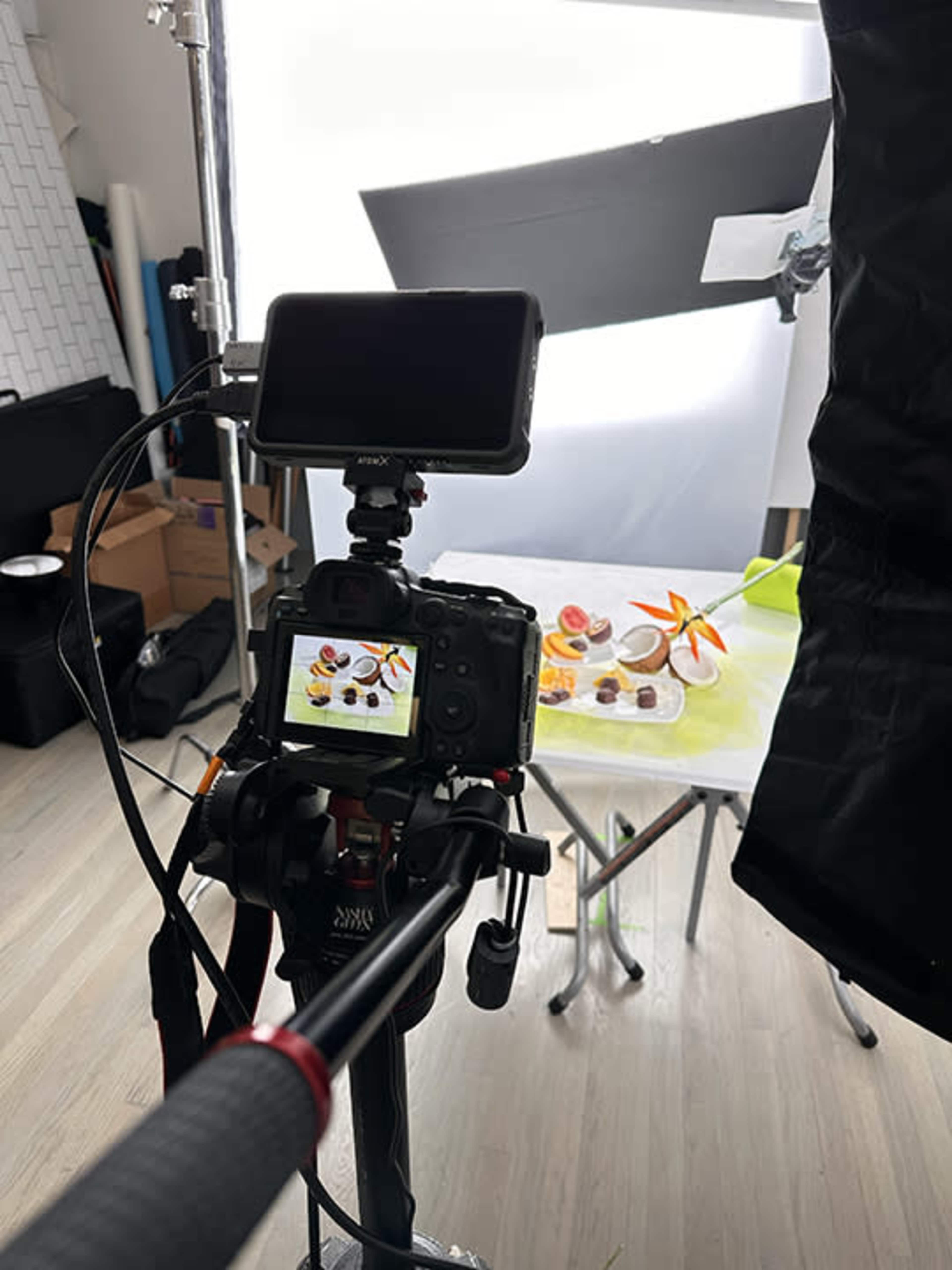 A camera is set up on a tripod, aimed at a table displaying various fruits and desserts in a photography studio.