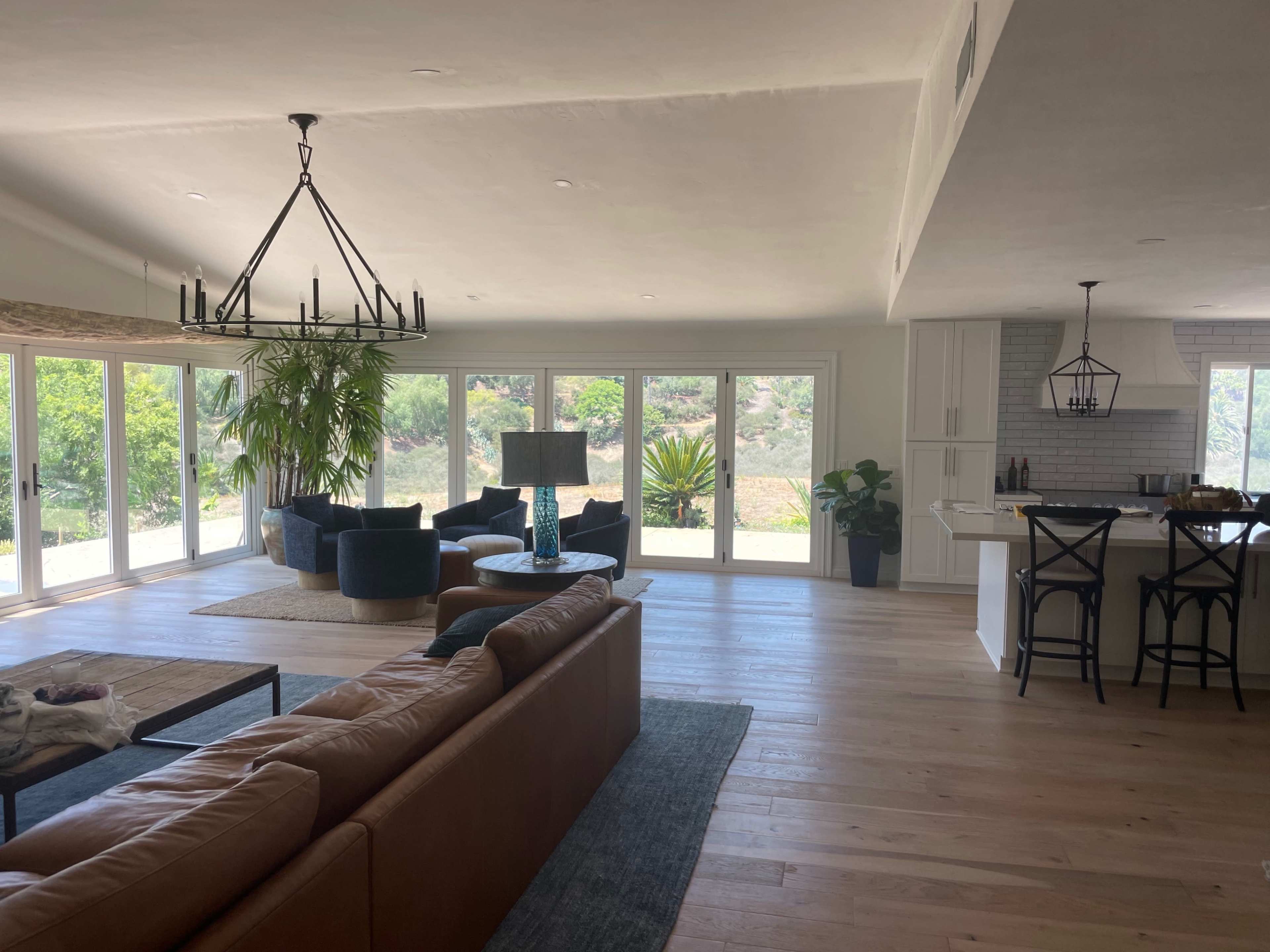 A modern living room with large windows overlooking greenery, featuring a brown leather sofa, a rug, two dark chairs, and a kitchen area in the background.