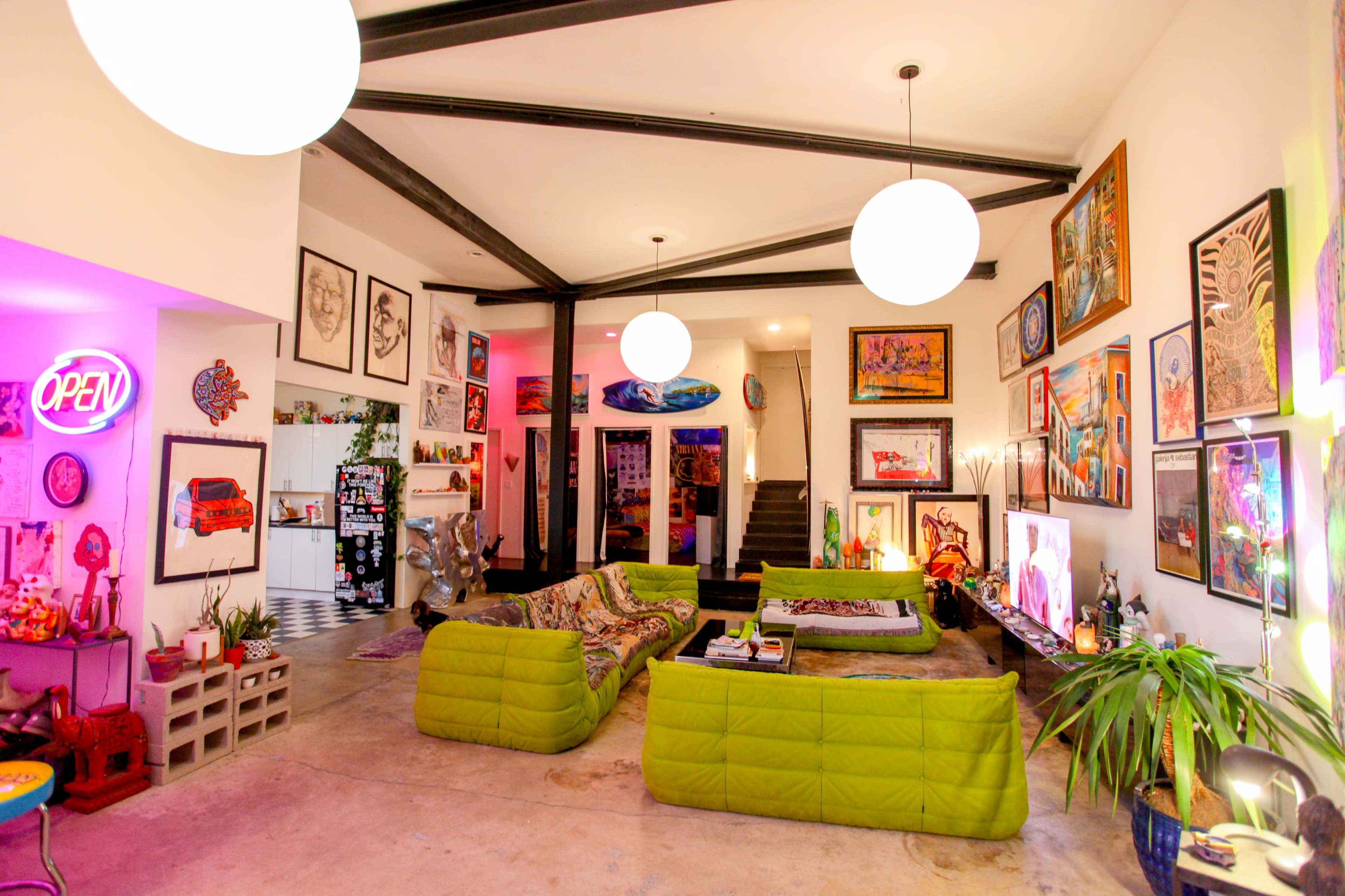 The image shows a brightly lit living room with green sofas arranged in a U-shape, adorned with various artworks on the walls, and a staircase visible in the background.