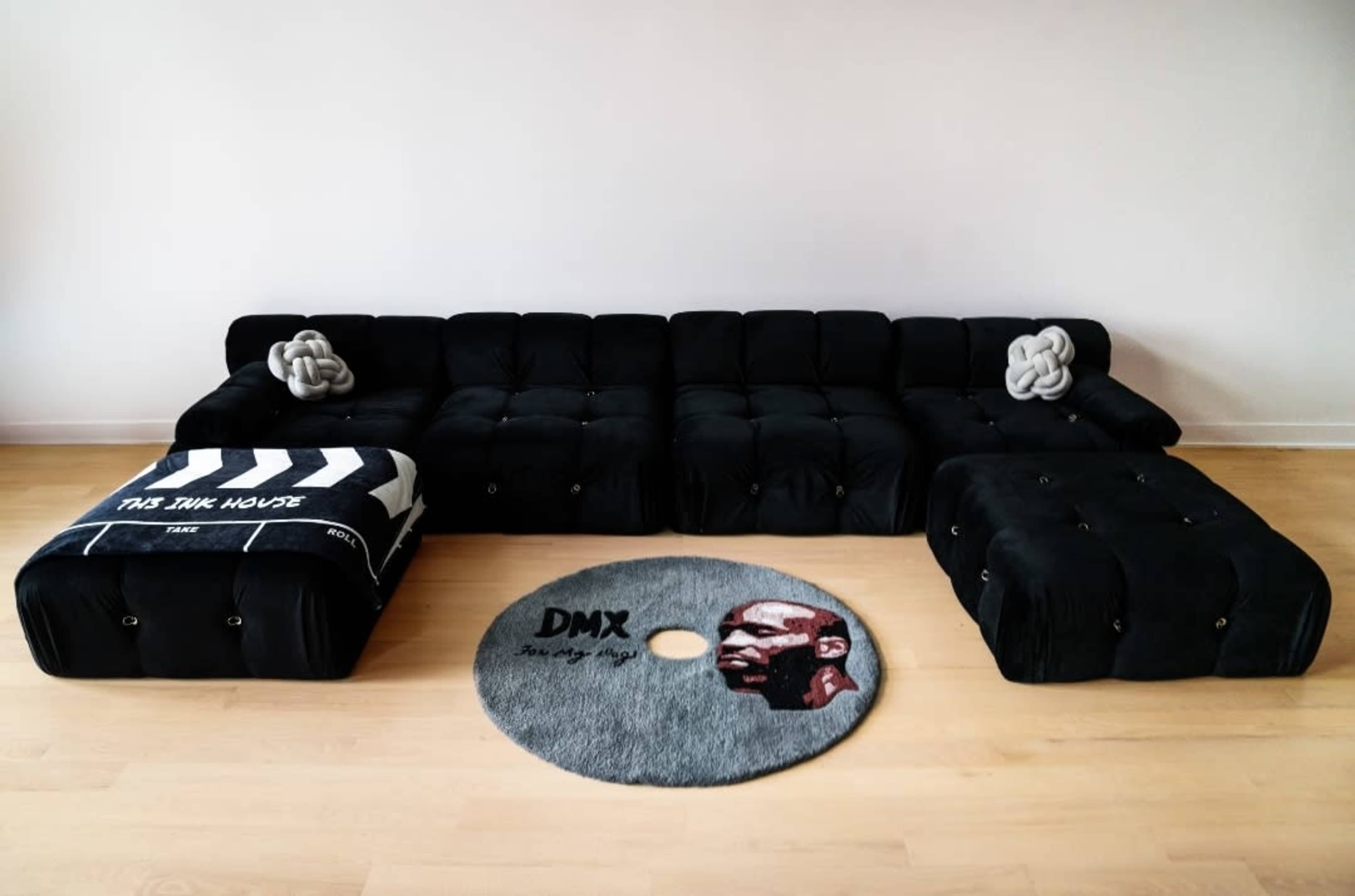A contemporary black sectional sofa arranged in a minimalist room with a round gray rug displaying a graphic of a man and the text "DMX."