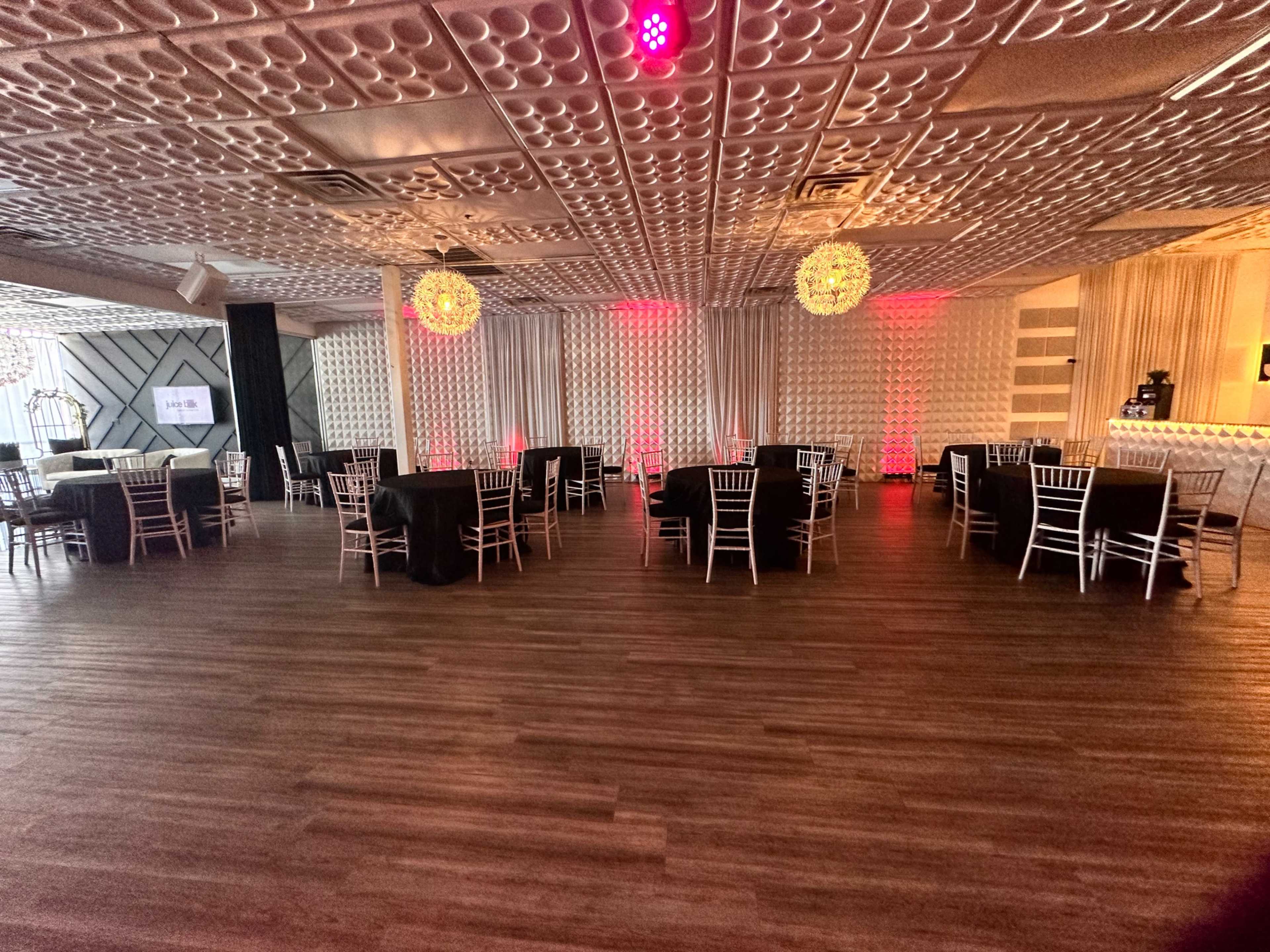 A spacious event venue set up with round tables covered in black tablecloths and elegant chairs, featuring decorative lighting and a bar area in the background.