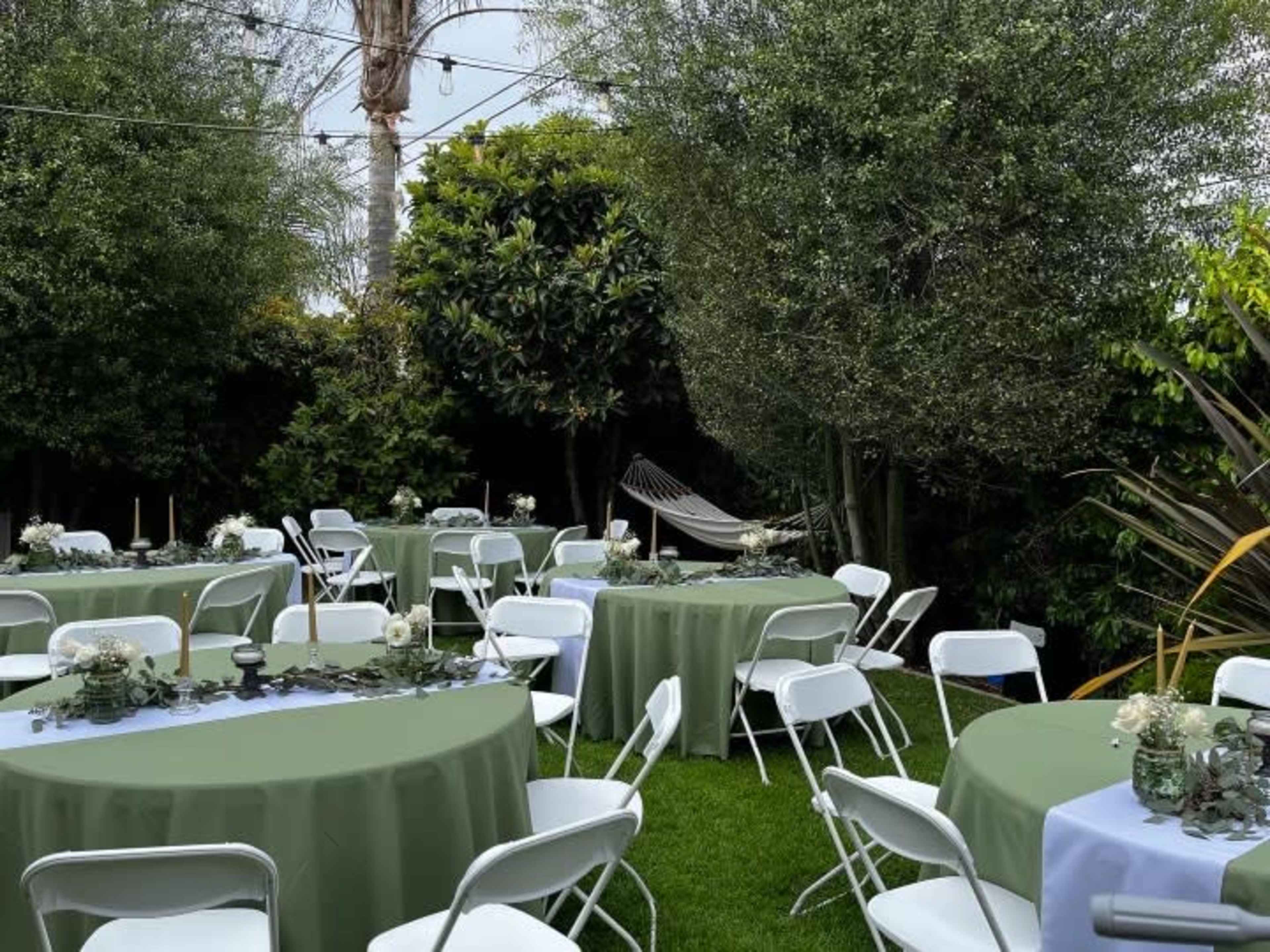 A garden setting with round tables covered in green tablecloths, surrounded by trees and a hammock in the background.