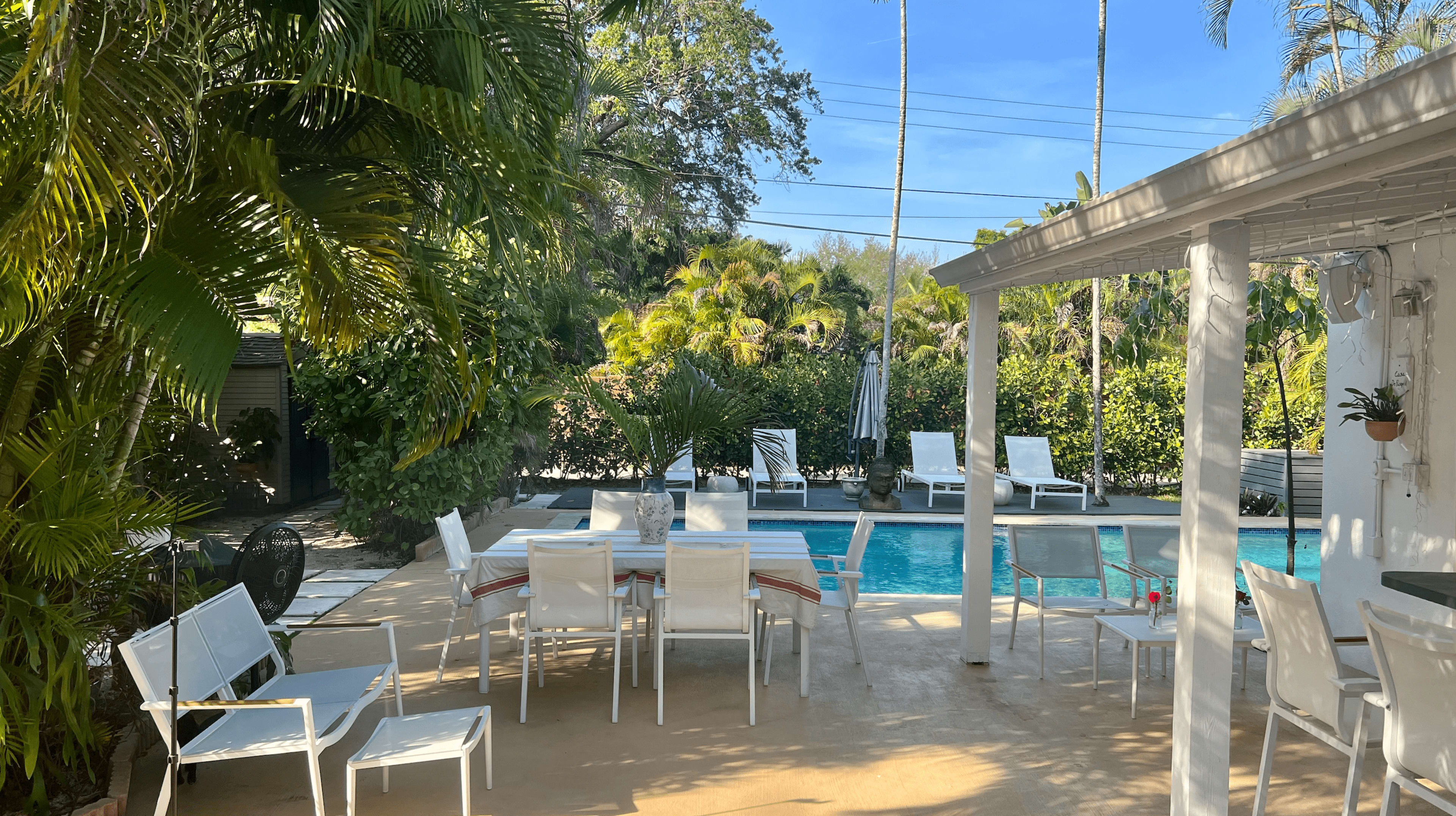 A patio area with white outdoor furniture surrounds a swimming pool, framed by palm trees and lush greenery.