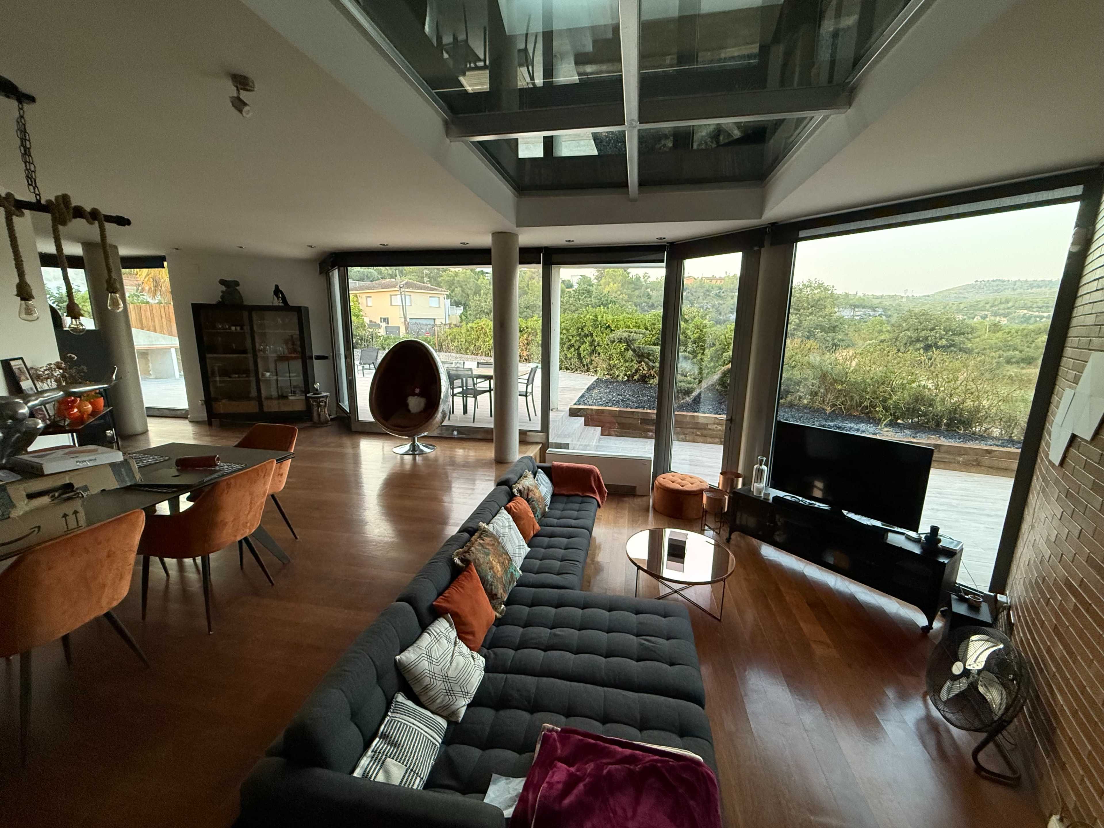 The image shows a modern living space with a large sectional sofa, a dining area, and floor-to-ceiling windows offering a view of a landscaped outdoor area.