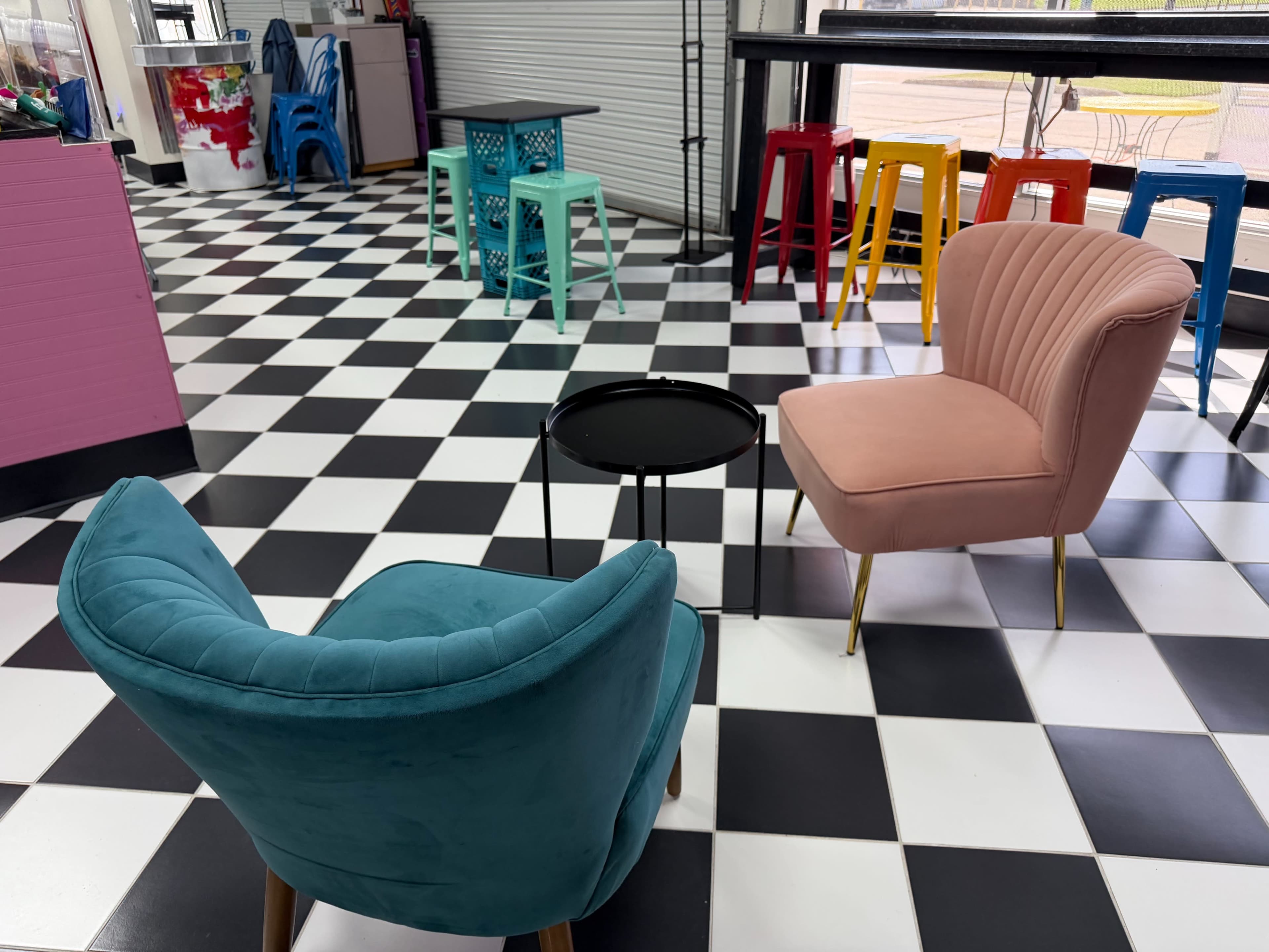 The image shows a seating area with two chairs, one teal and one pink, positioned around a small black table on a black and white checkered floor.
