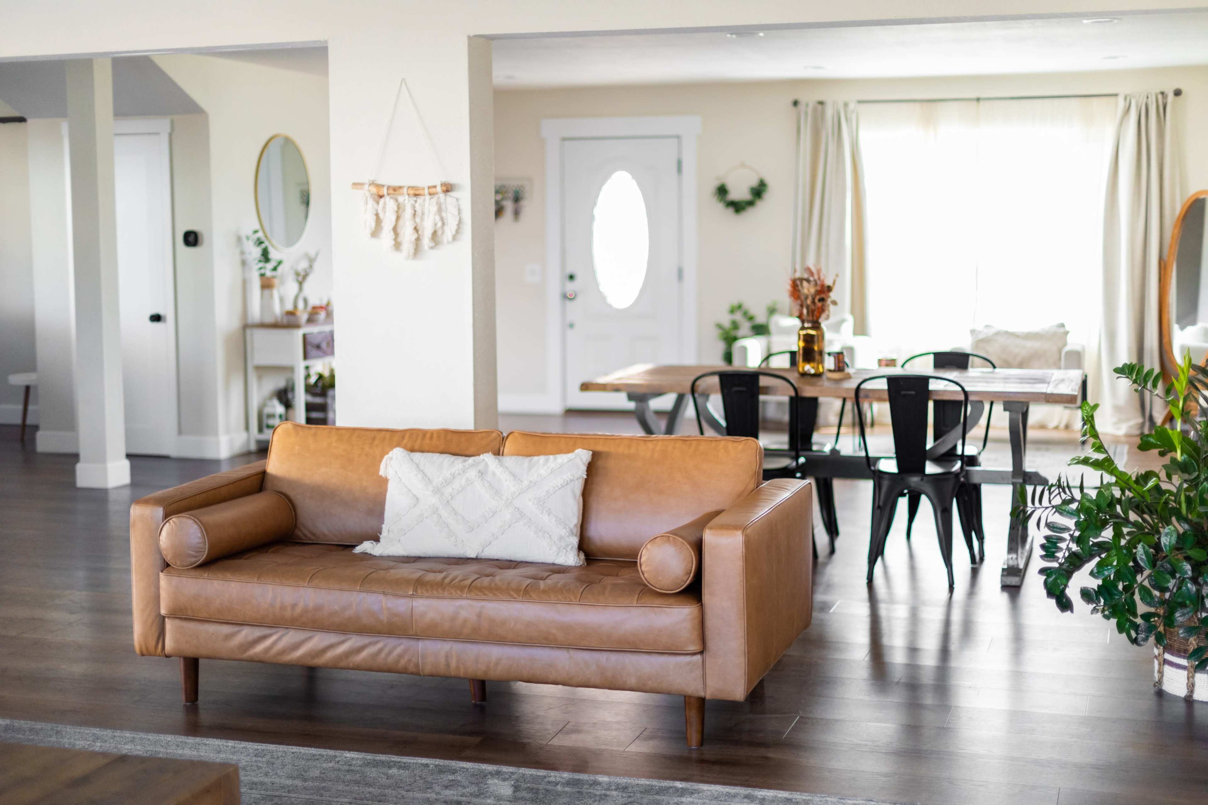 A leather sofa with a decorative pillow is positioned in a light-filled living area that opens to a dining space with a wooden table and chairs.