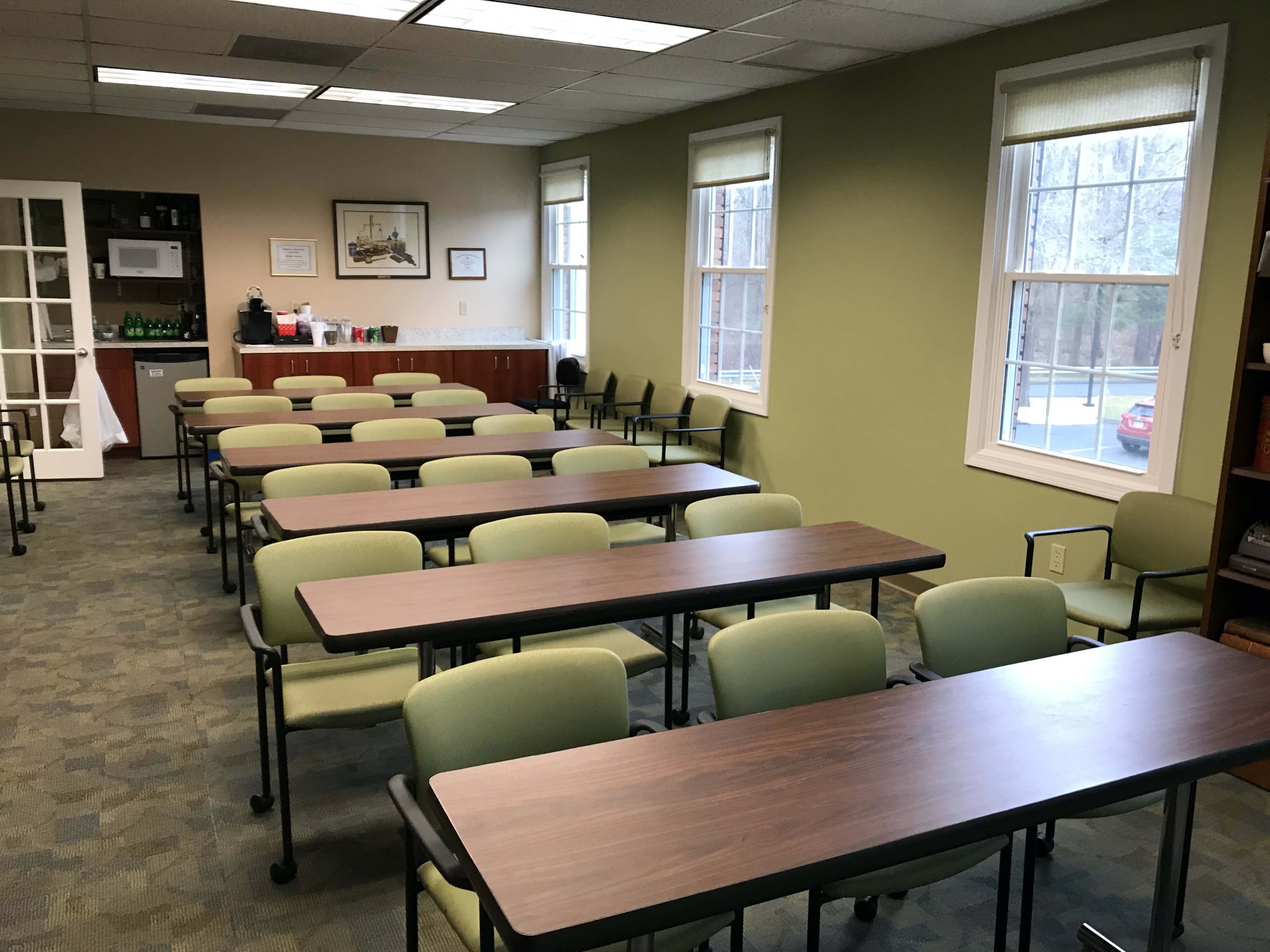 A classroom setting with several rows of green chairs and tables, along with a kitchenette and windows providing natural light.