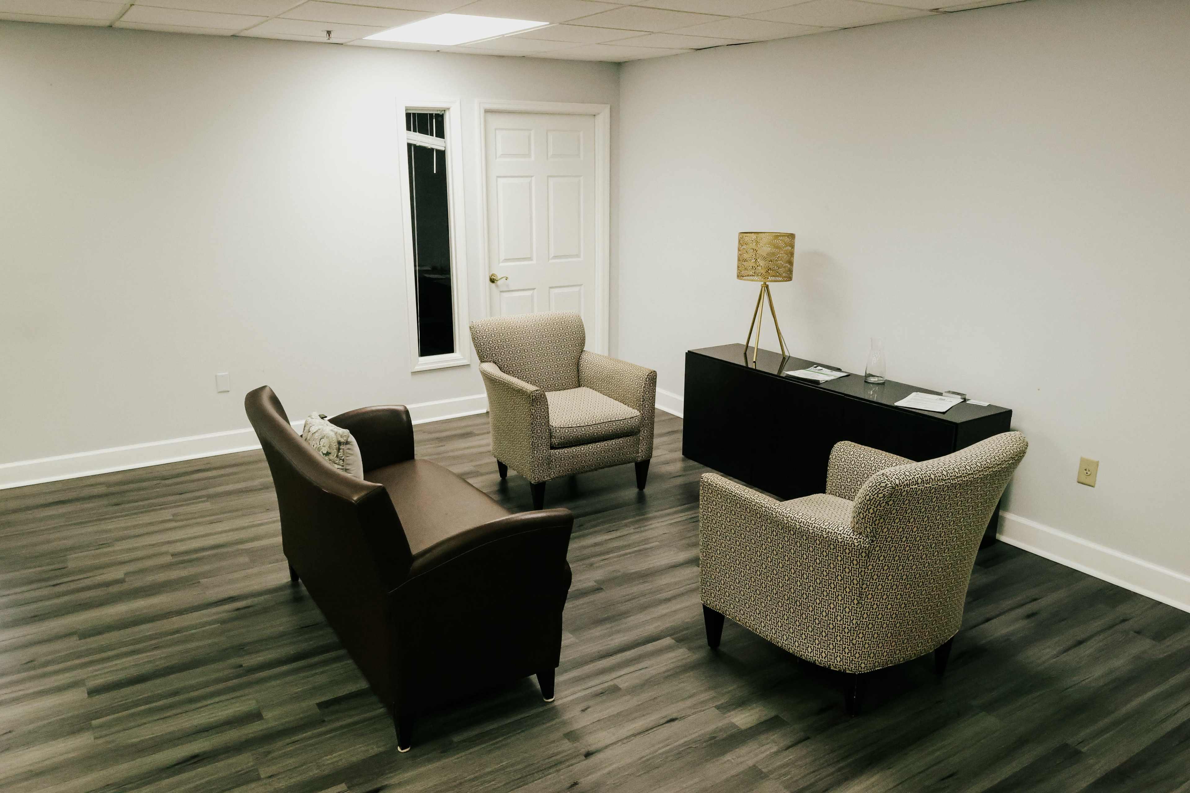 A small, minimalist office space featuring two armchairs and a loveseat arranged around a black desk with a lamp and some paperwork.