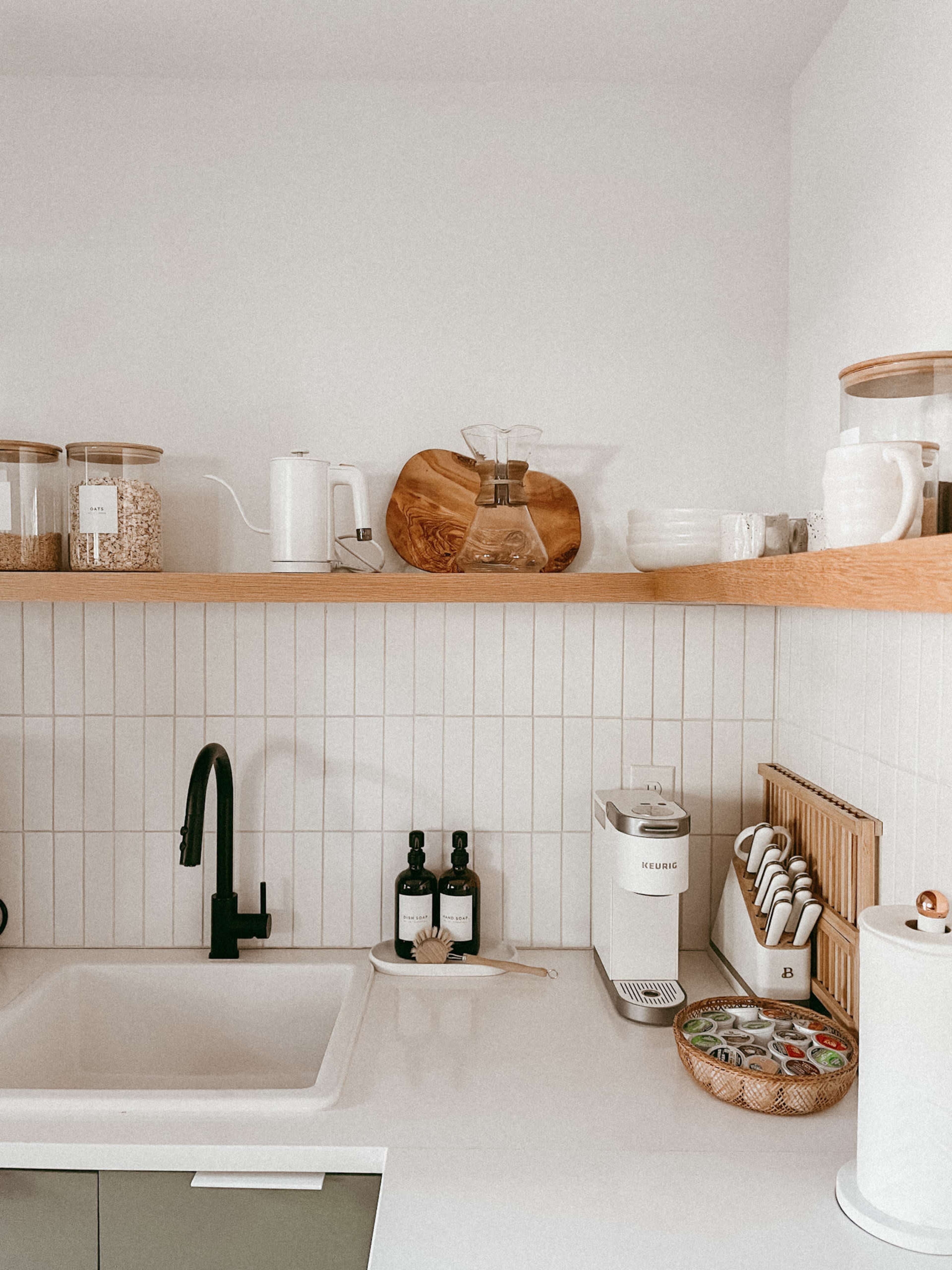 A modern kitchen features a white countertop with a sink, wooden shelves displaying jars and utensils, and a coffee maker.