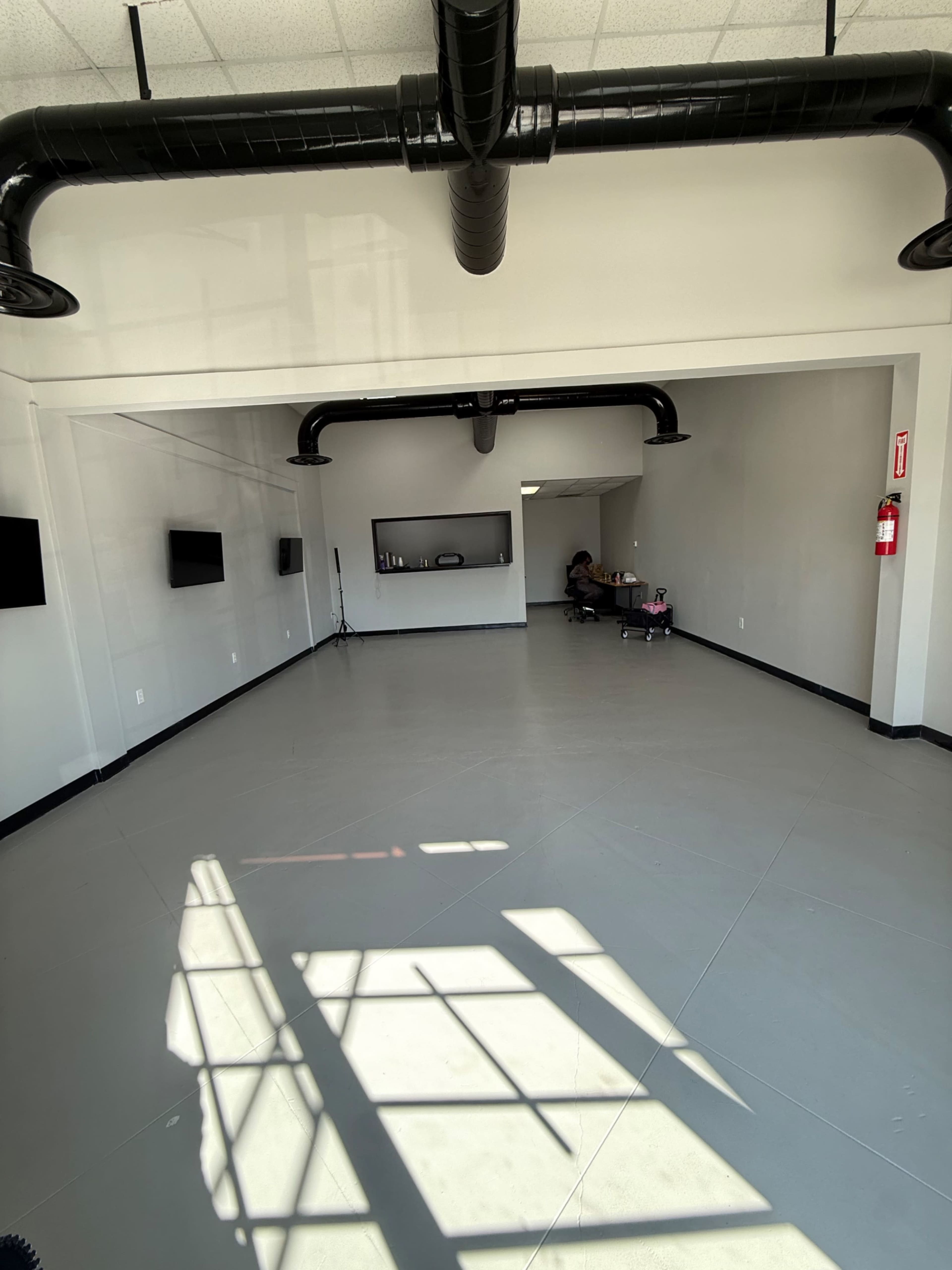 The image shows a large, empty room with grey flooring, white walls, and a few mounted flat-screen displays, along with visible black air ducts overhead.