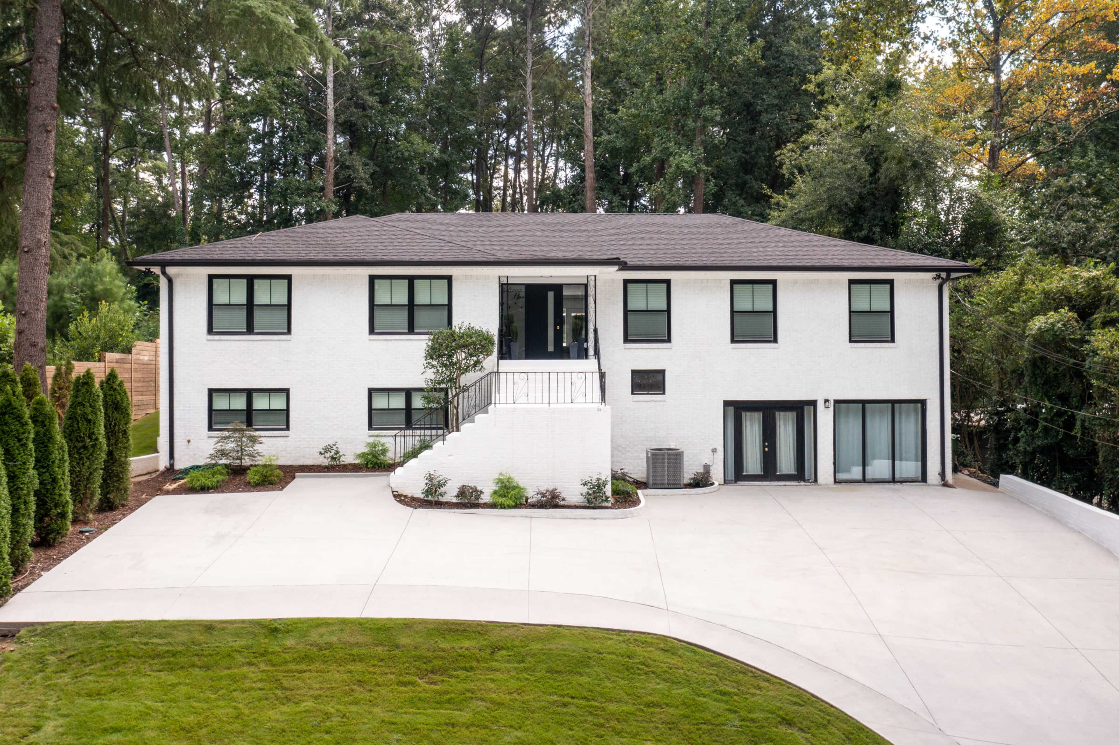 A modern two-story white house is surrounded by greenery with a large concrete driveway leading up to it.