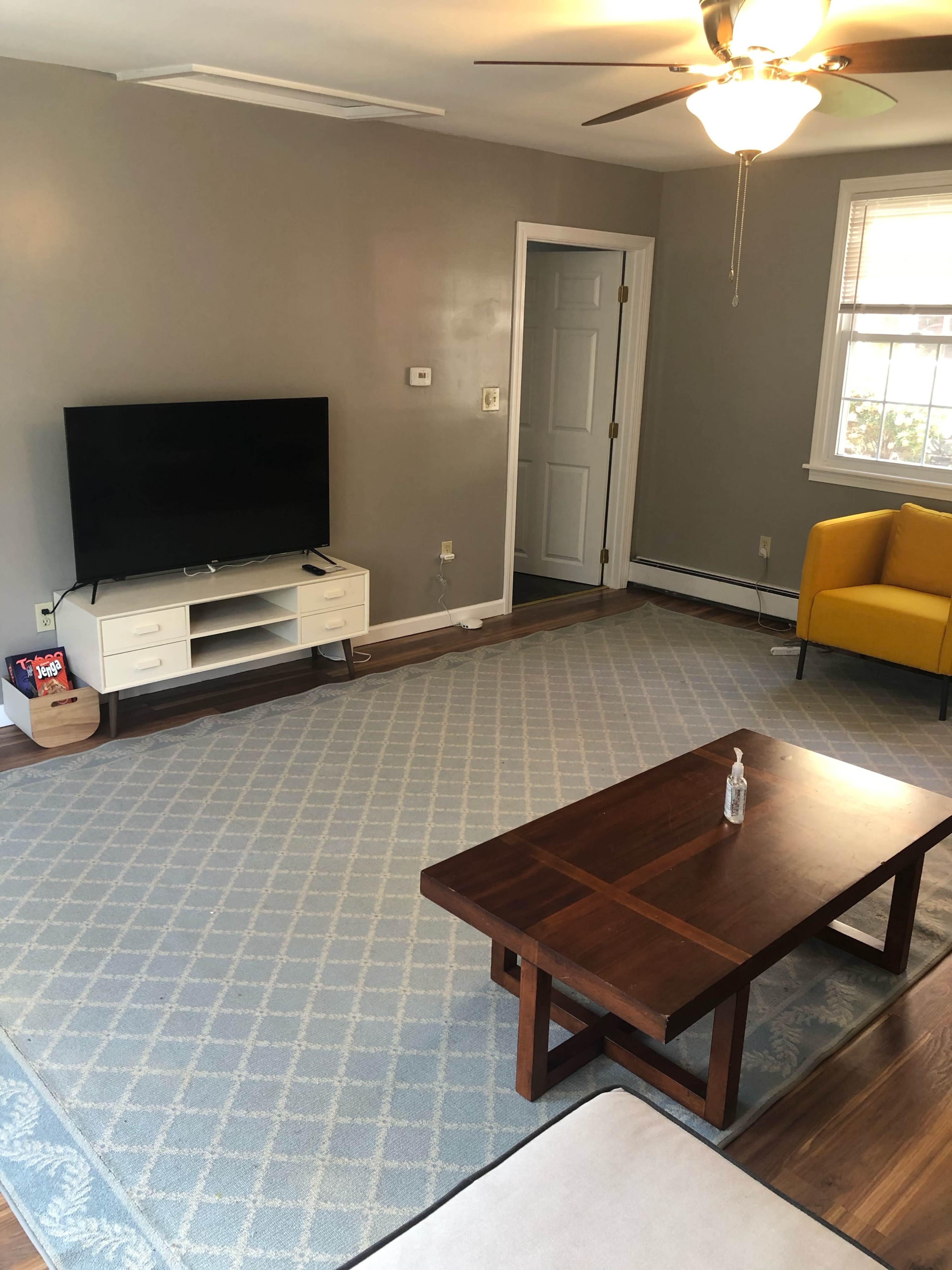 The living room features a television on a white stand, a wooden coffee table, a yellow chair, and a light blue area rug on a hardwood floor.