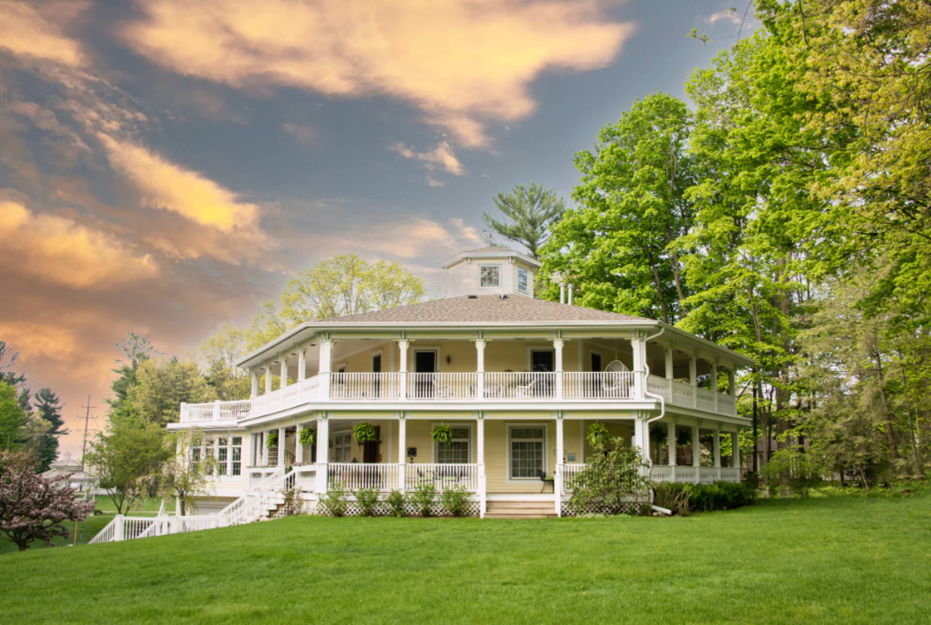 A large, two-story white house with a circular design, multiple balconies, and surrounded by greenery under a cloudy sky.
