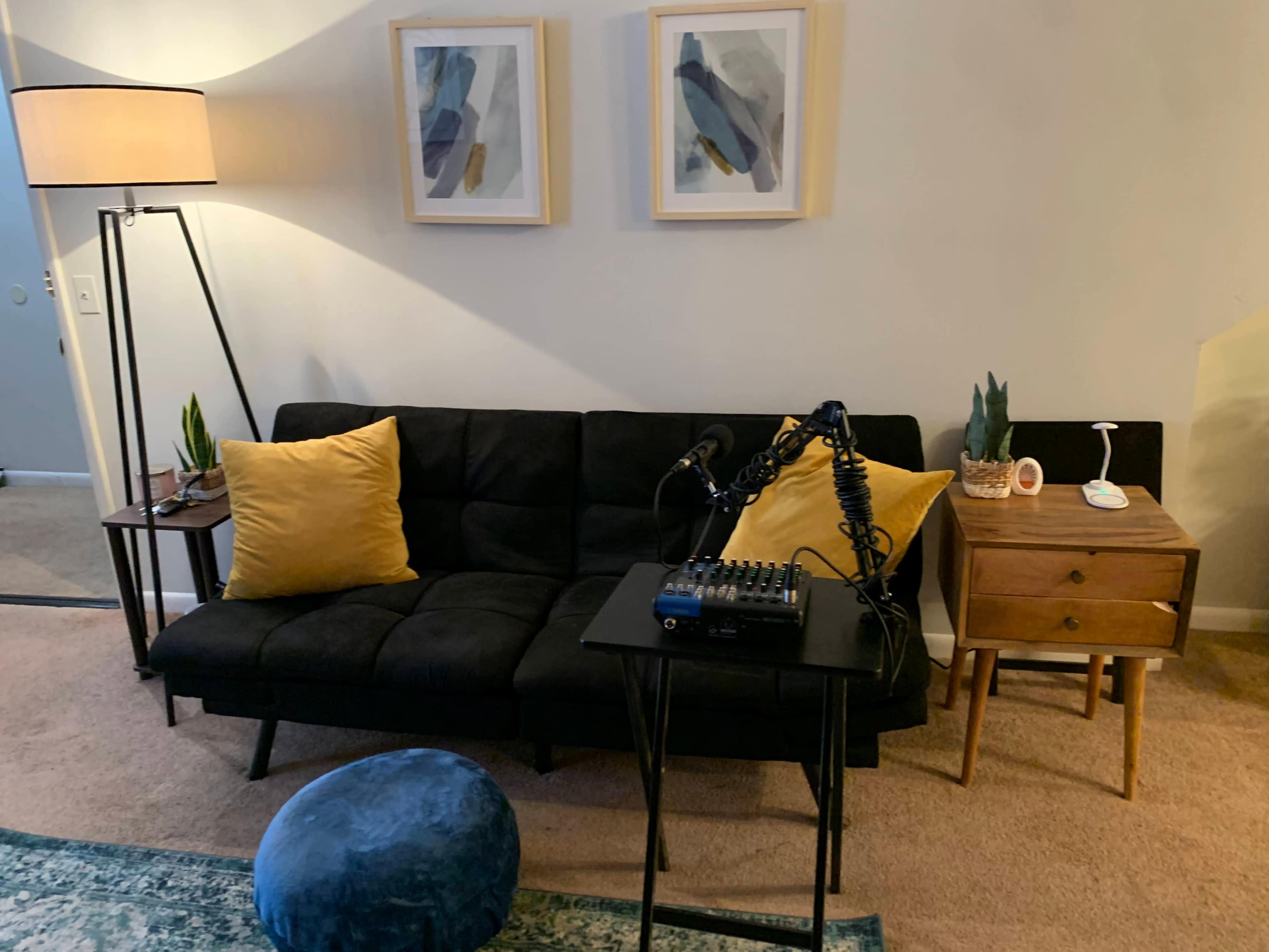 The room features a black couch with yellow accent pillows, a lamp, a small table with audio equipment, and framed artwork on the wall.