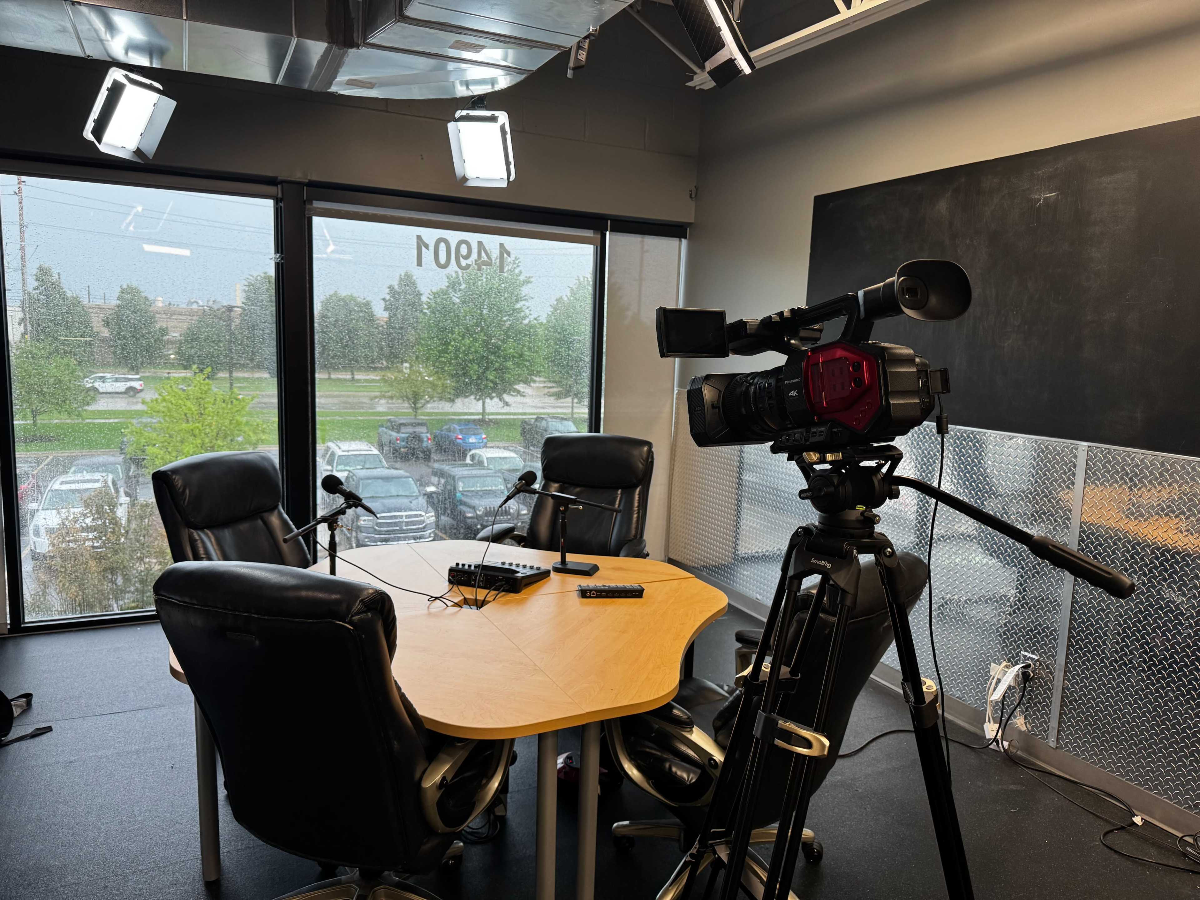 The image shows a video recording setup in a studio with a wooden table, three black chairs, and a camera on a tripod facing the table, with large windows showing an outdoor view.