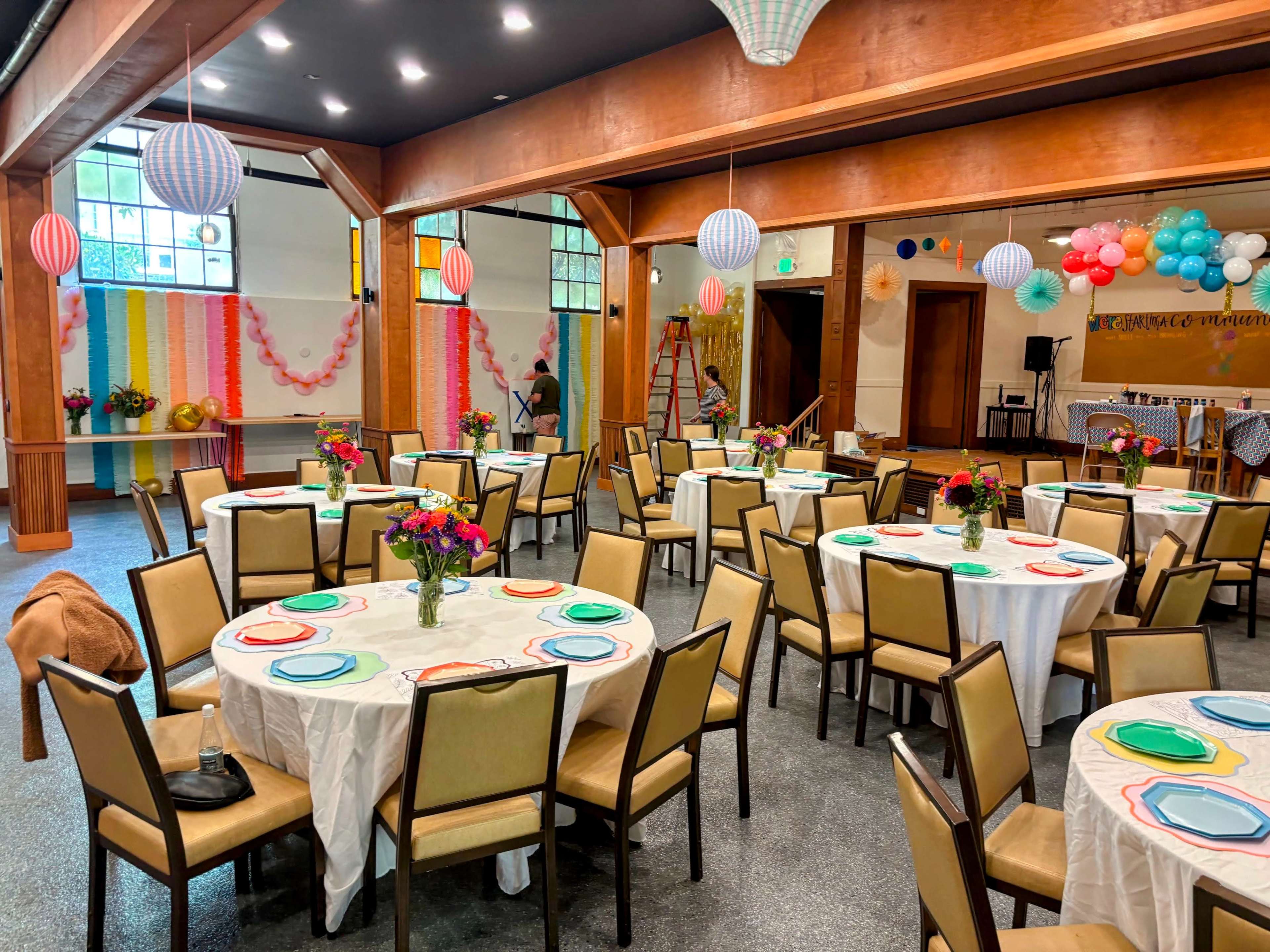 The image shows a reception hall decorated with colorful paper lanterns, flowers on each table, and a backdrop of bright streamers.