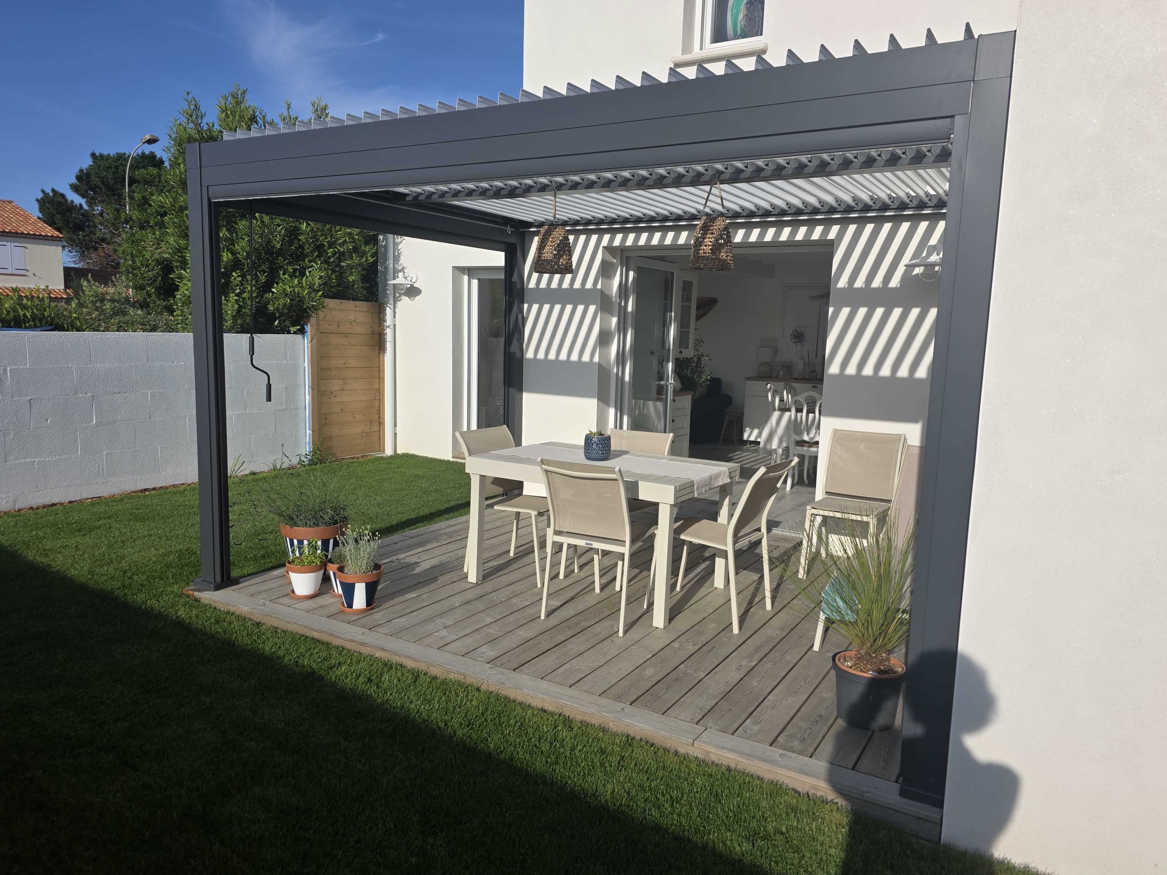 An outdoor patio features a wooden deck with a table surrounded by chairs, framed by a modern pergola and bordered by a green lawn.