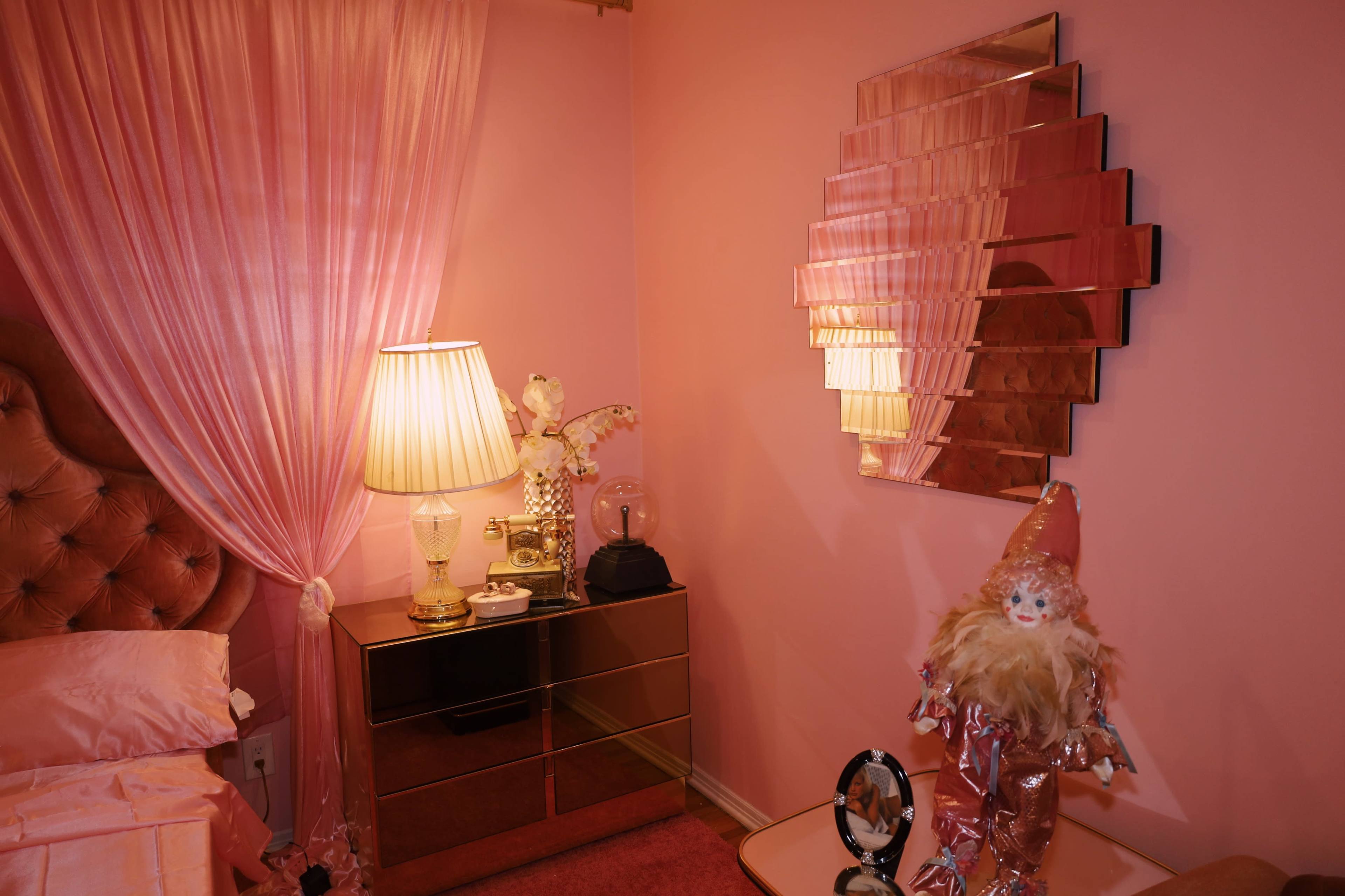 The image shows a pink-themed bedroom featuring a tufted headboard, a bedside lamp, a mirrored wall decoration, and a decorative clown figurine.