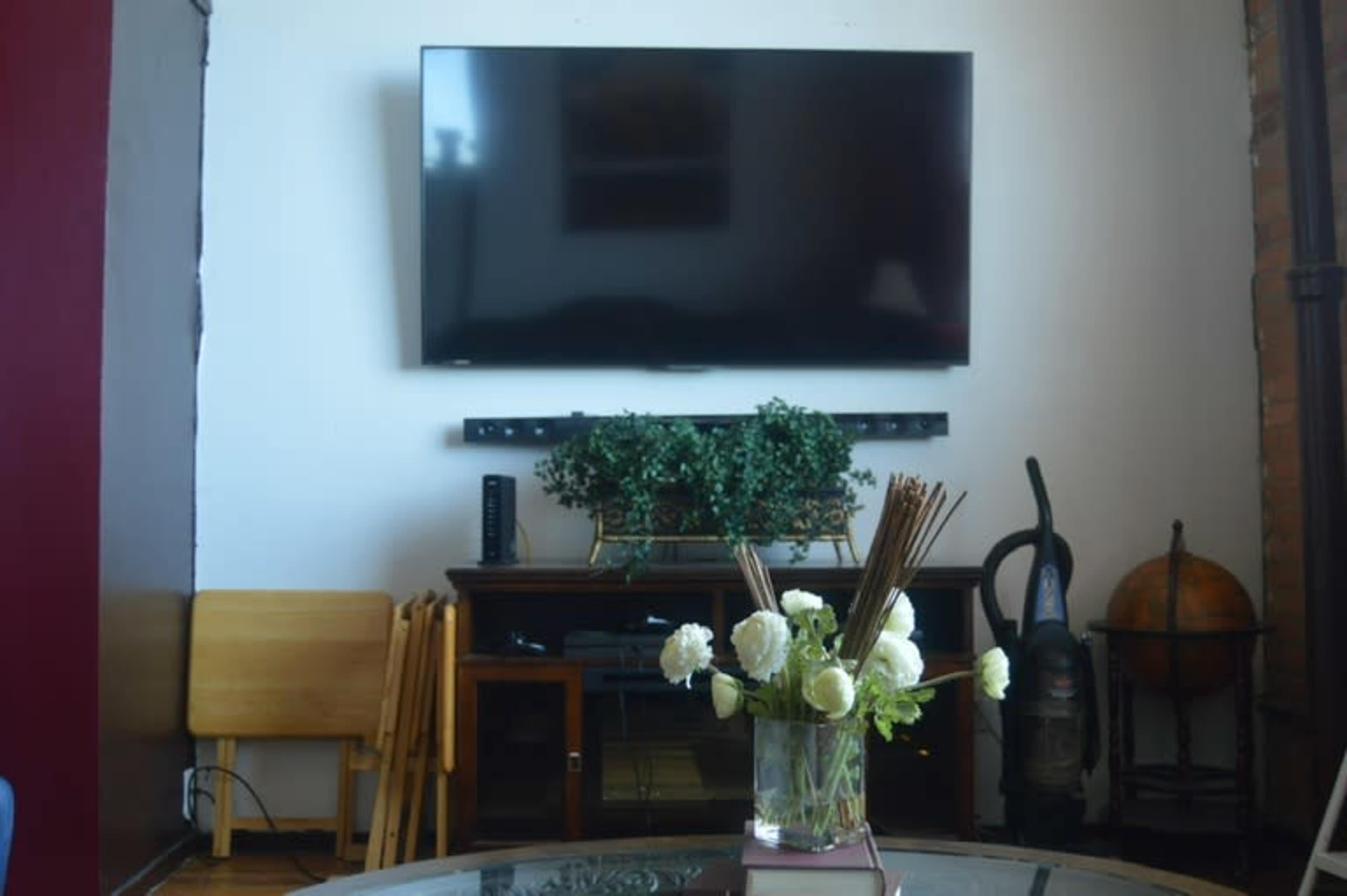 A living room with a mounted TV, a wooden table with a vase of flowers, and a decorative plant on a cabinet.