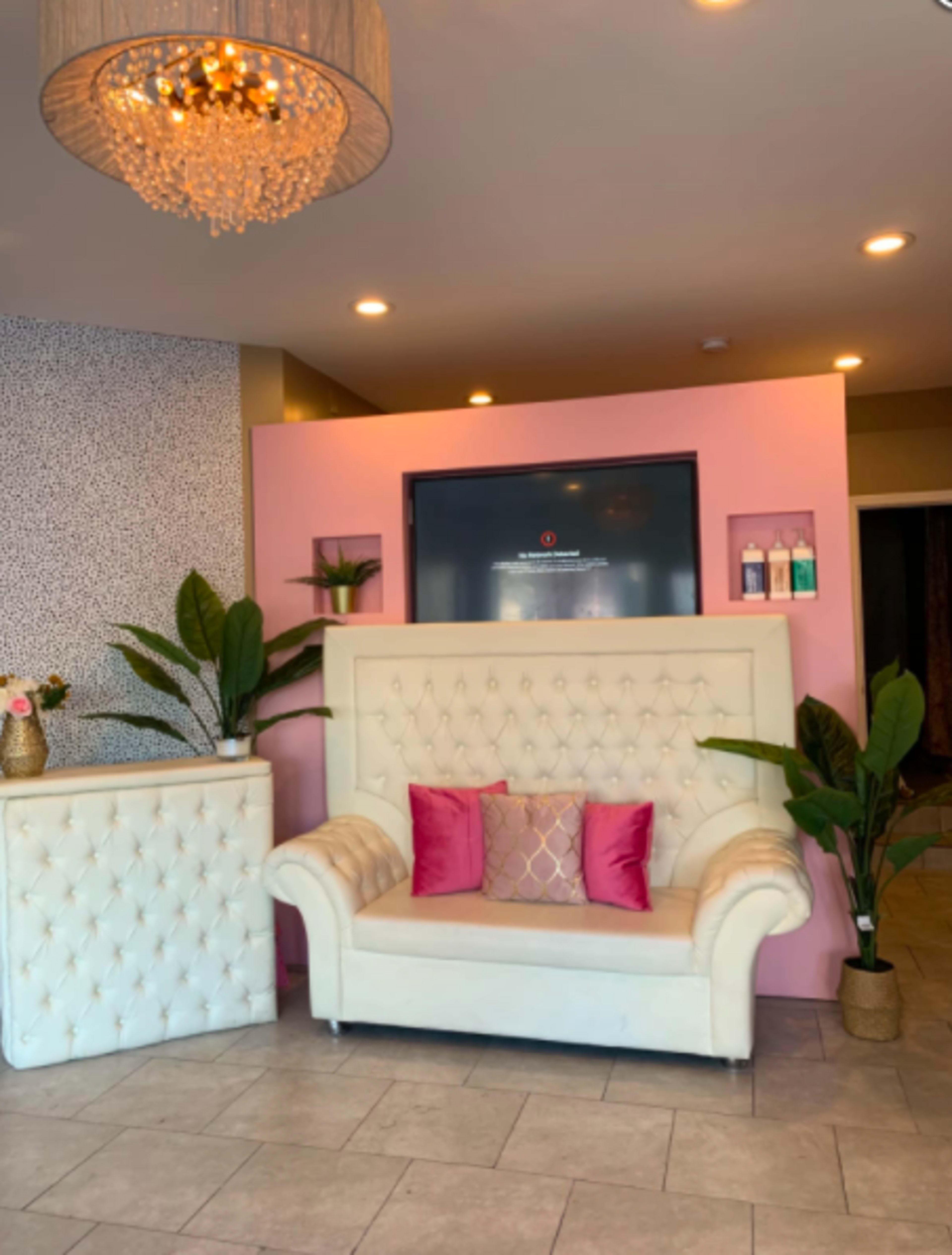 A modern reception area featuring a white tufted sofa, pink accent pillows, and decorative plants, with a large television mounted on a pink wall and a light fixture overhead.