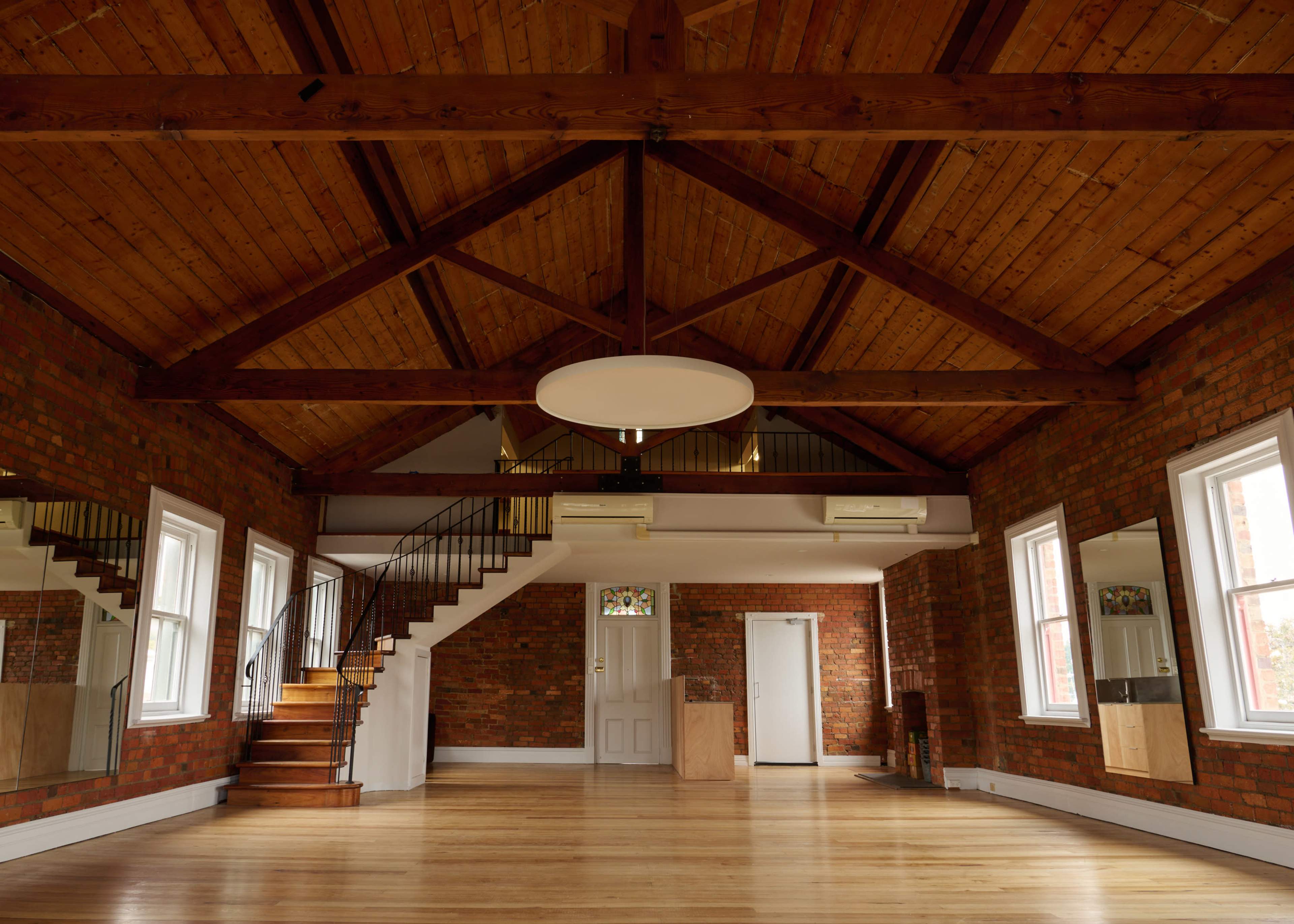 The image shows a spacious room with wooden floors, exposed brick walls, and a high ceiling featuring wooden beams, along with a staircase in the background.