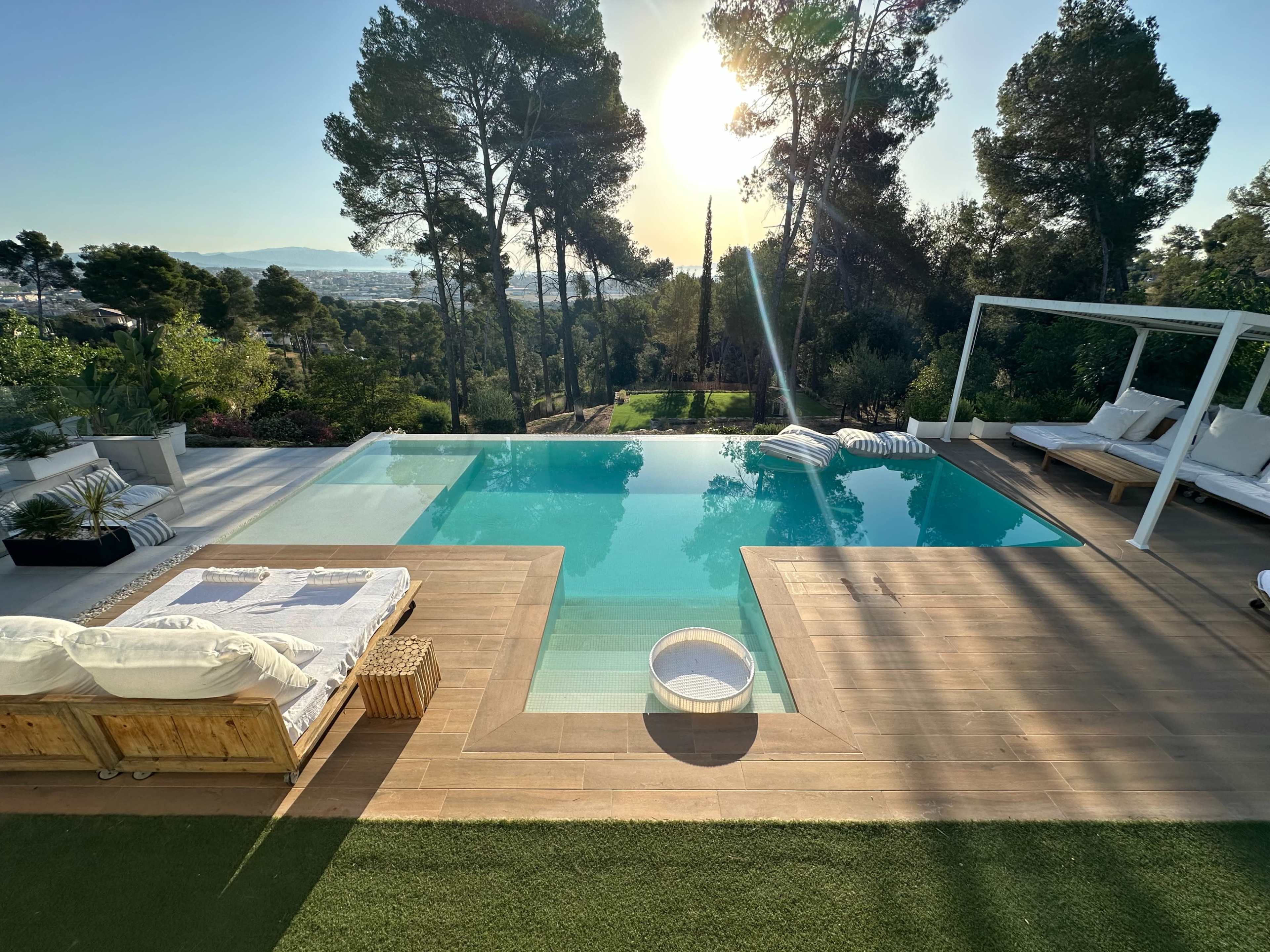 A modern pool area features an infinity pool surrounded by wooden decking, lounge chairs, and lush greenery under a clear sky with the sun setting in the background.