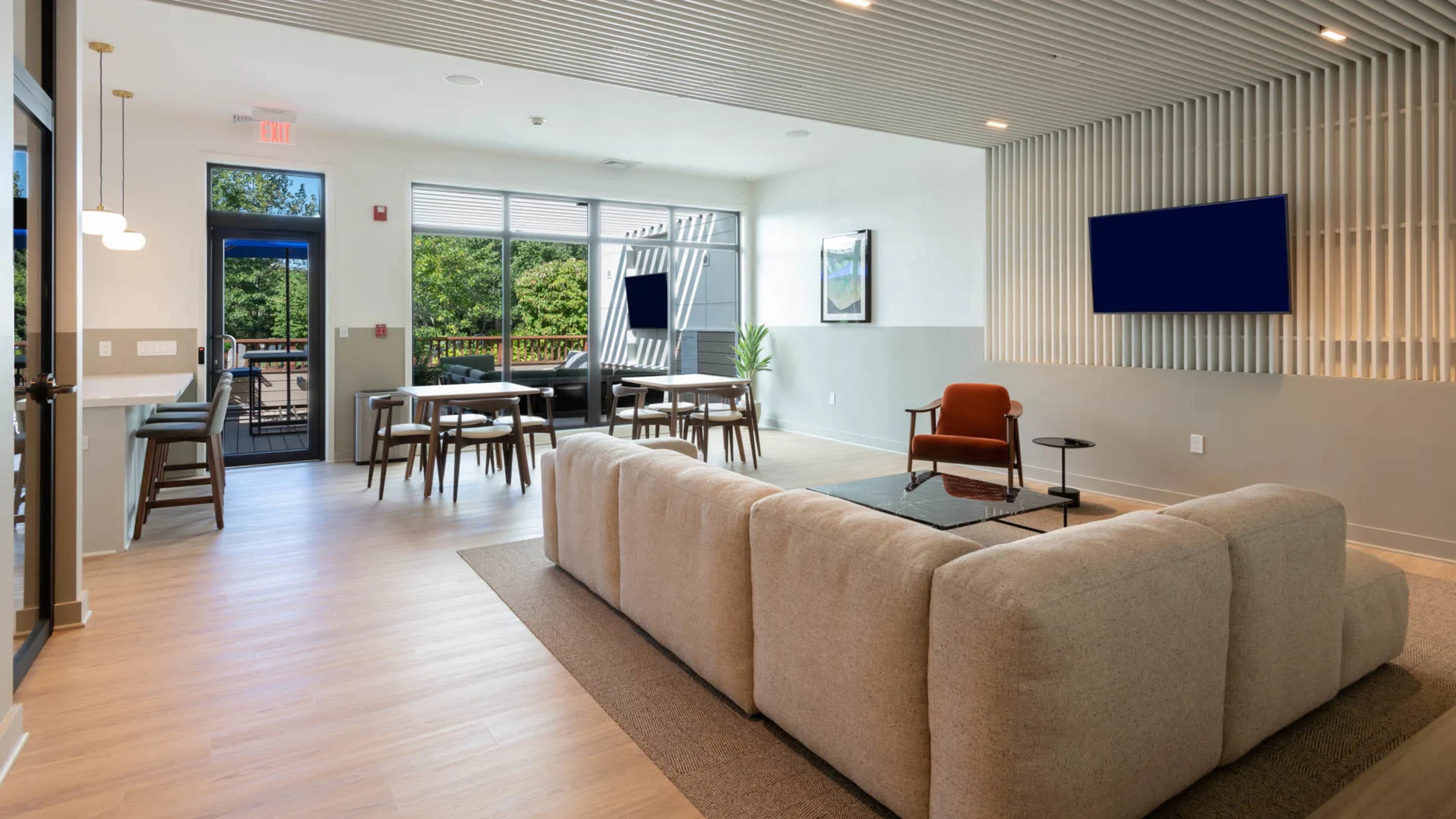 The image shows a modern lounge area featuring a large sectional sofa, a coffee table, and multiple seating arrangements, with a view of an outdoor space through large windows.