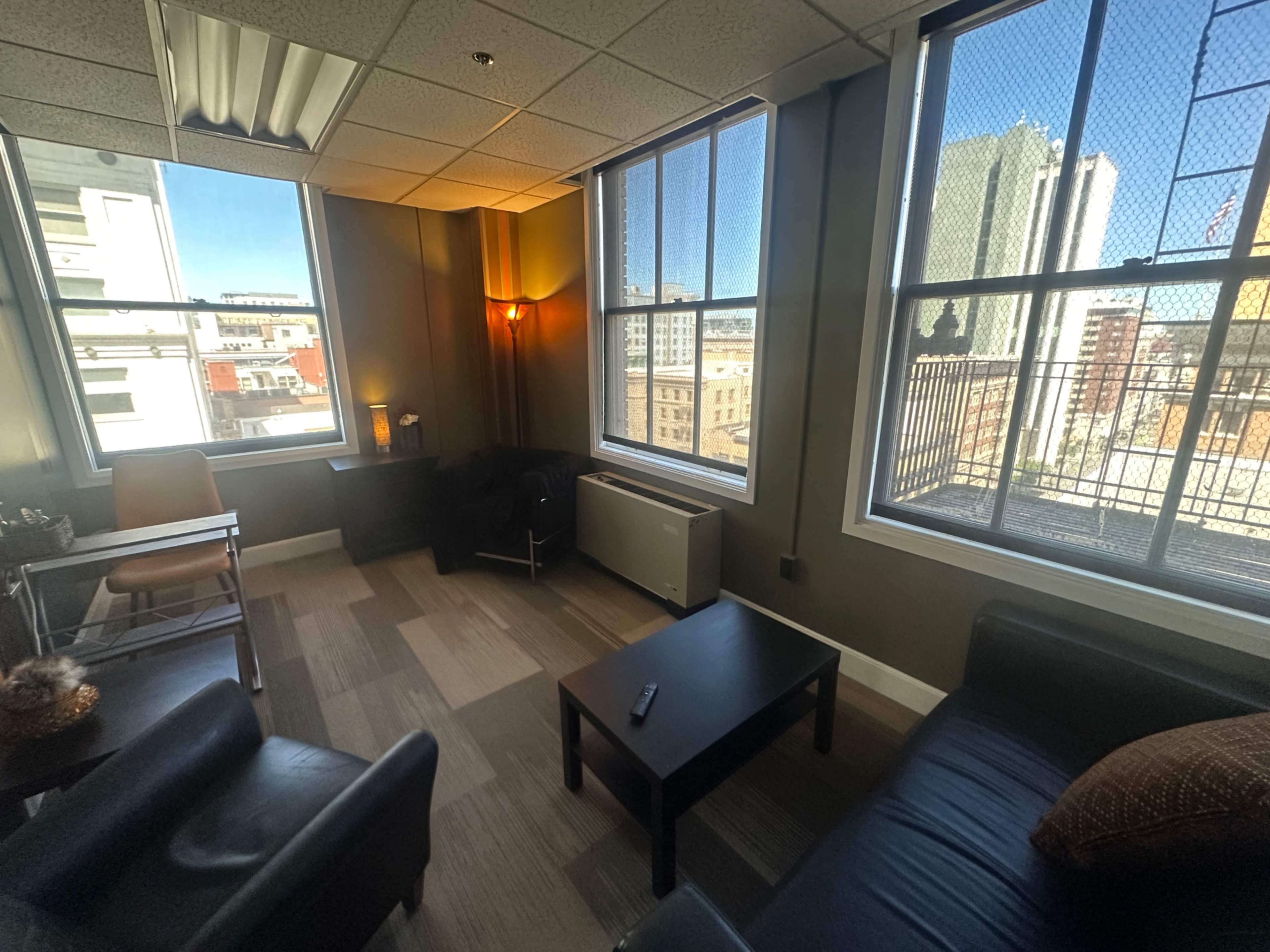 The image shows a corner office with large windows, a small table, black couches, and a warm light fixture.