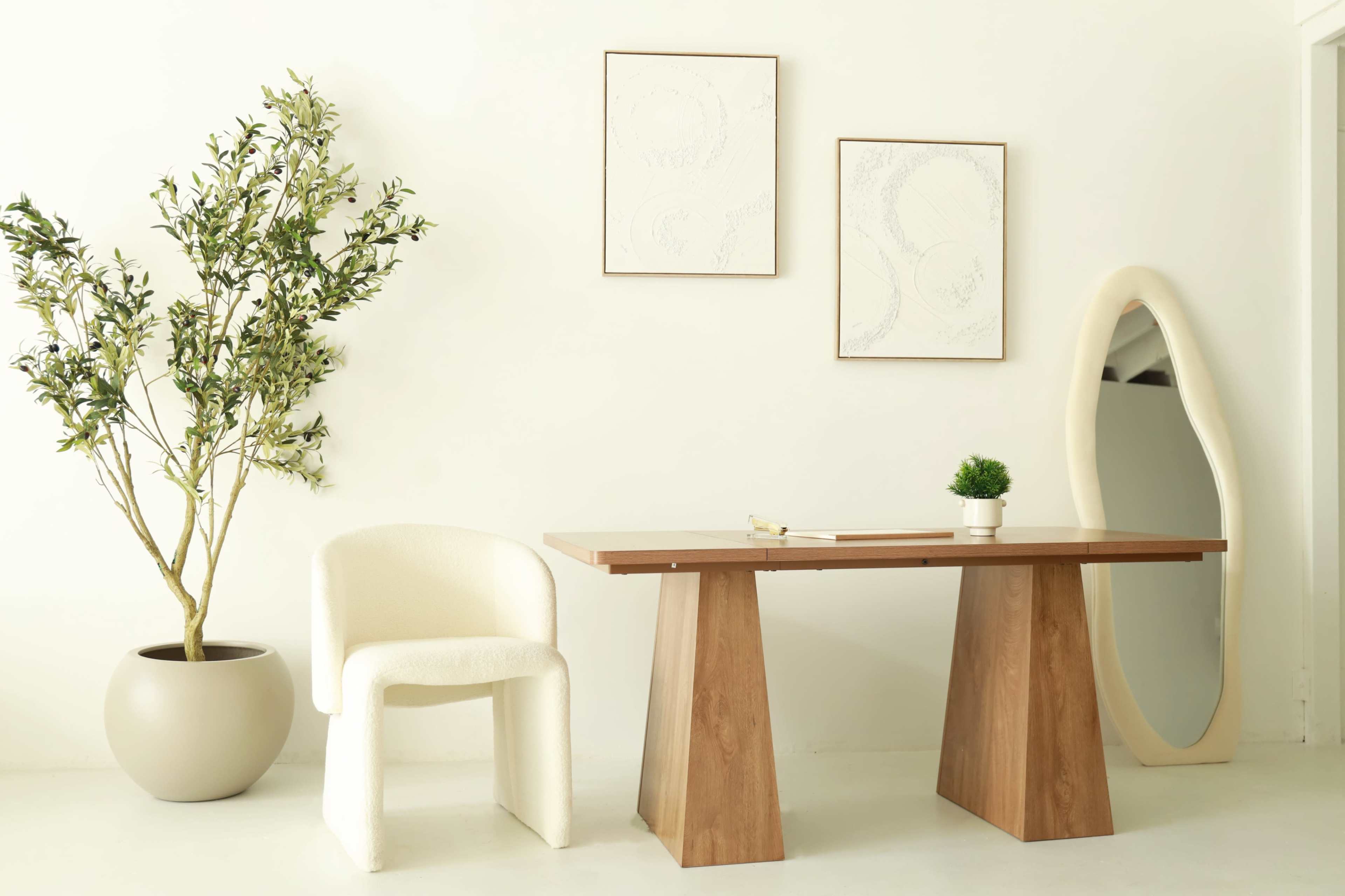 The image shows a minimalist workspace featuring a wooden table, a textured chair, a potted plant, and two framed artworks on a white wall.