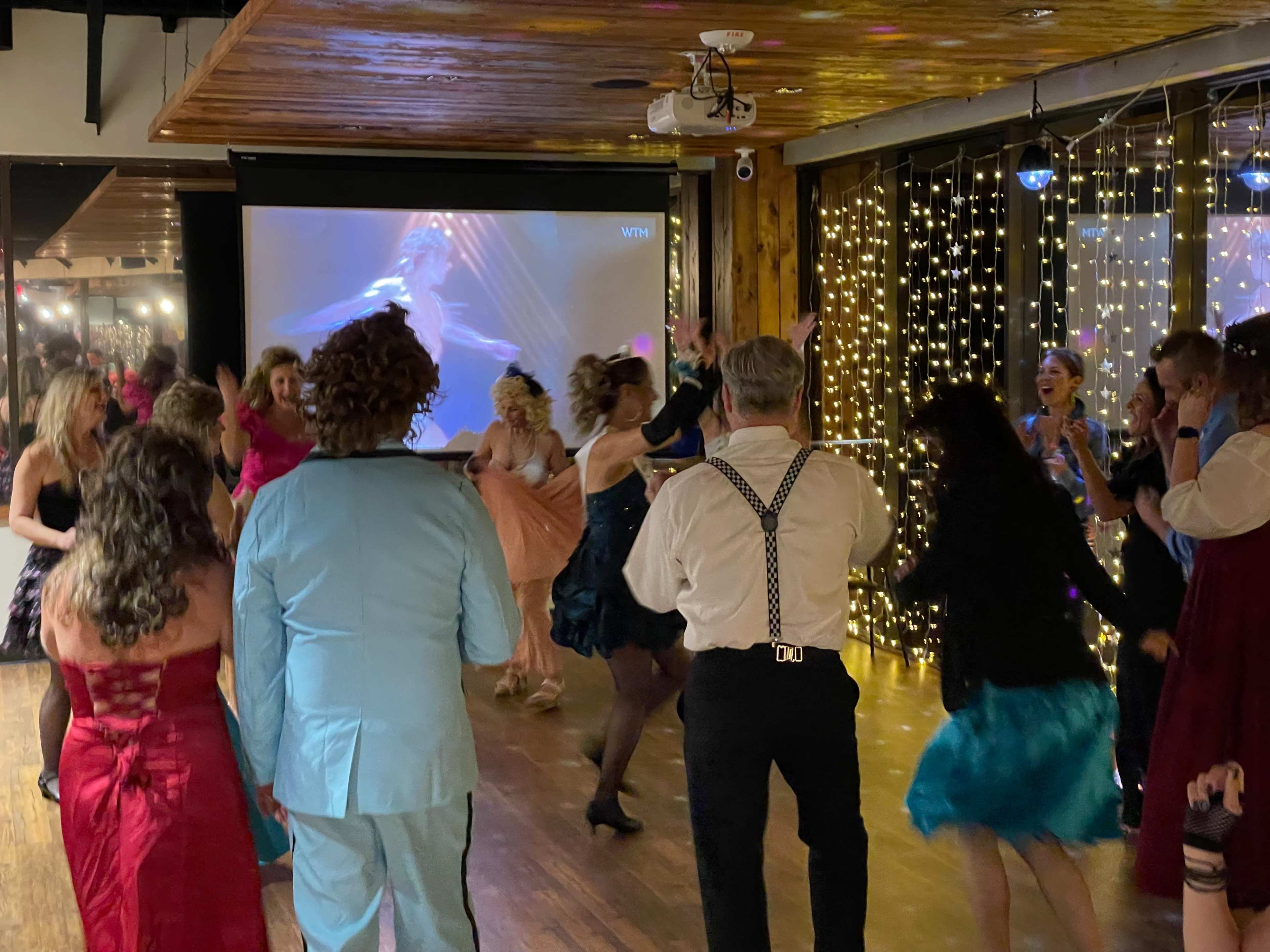 A lively group of people is dancing on a wooden floor in a decorated venue with twinkling lights and a projected image in the background.