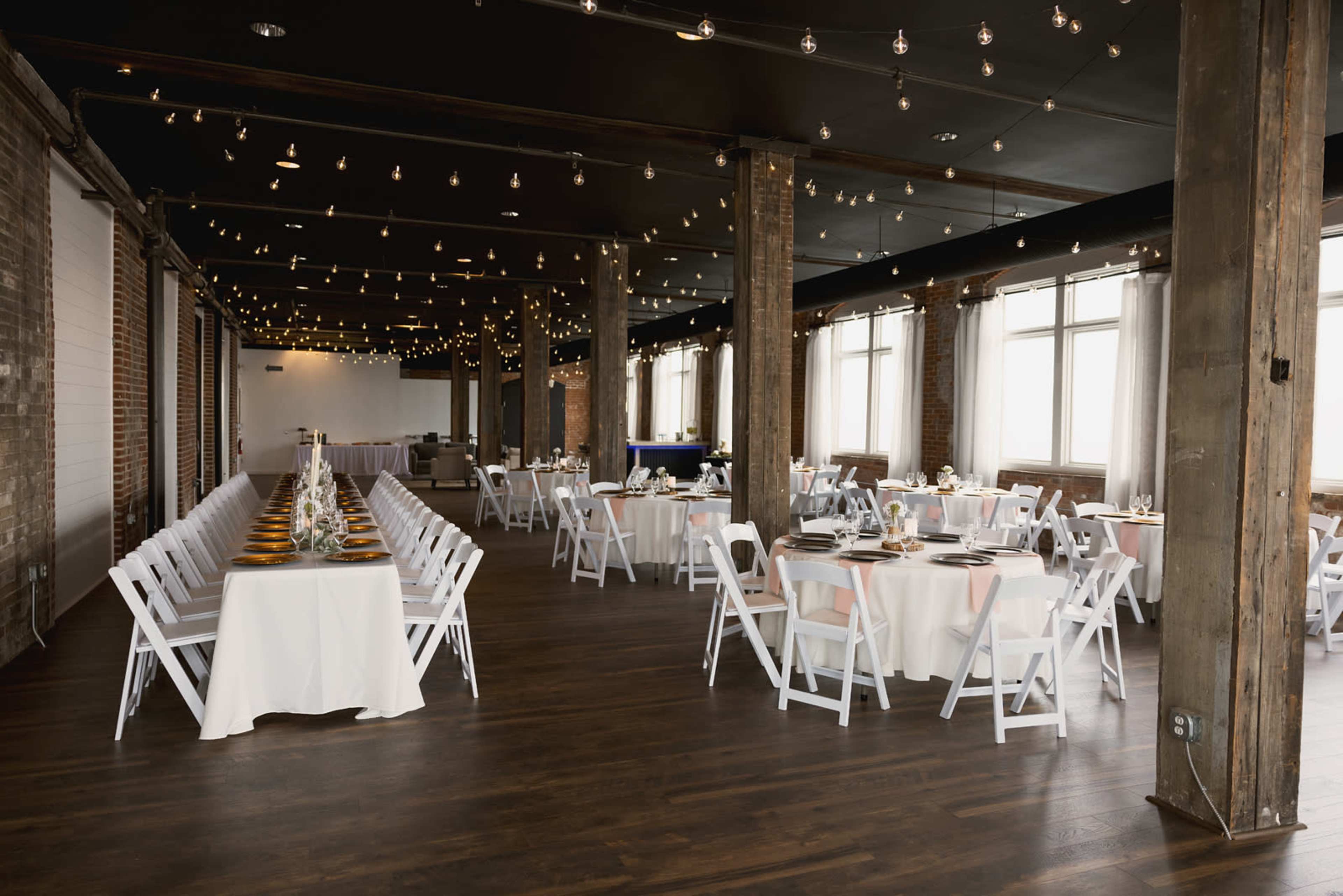 A spacious event hall features rows of elegantly set tables, adorned with white tablecloths and surrounded by exposed brick walls and hanging string lights.