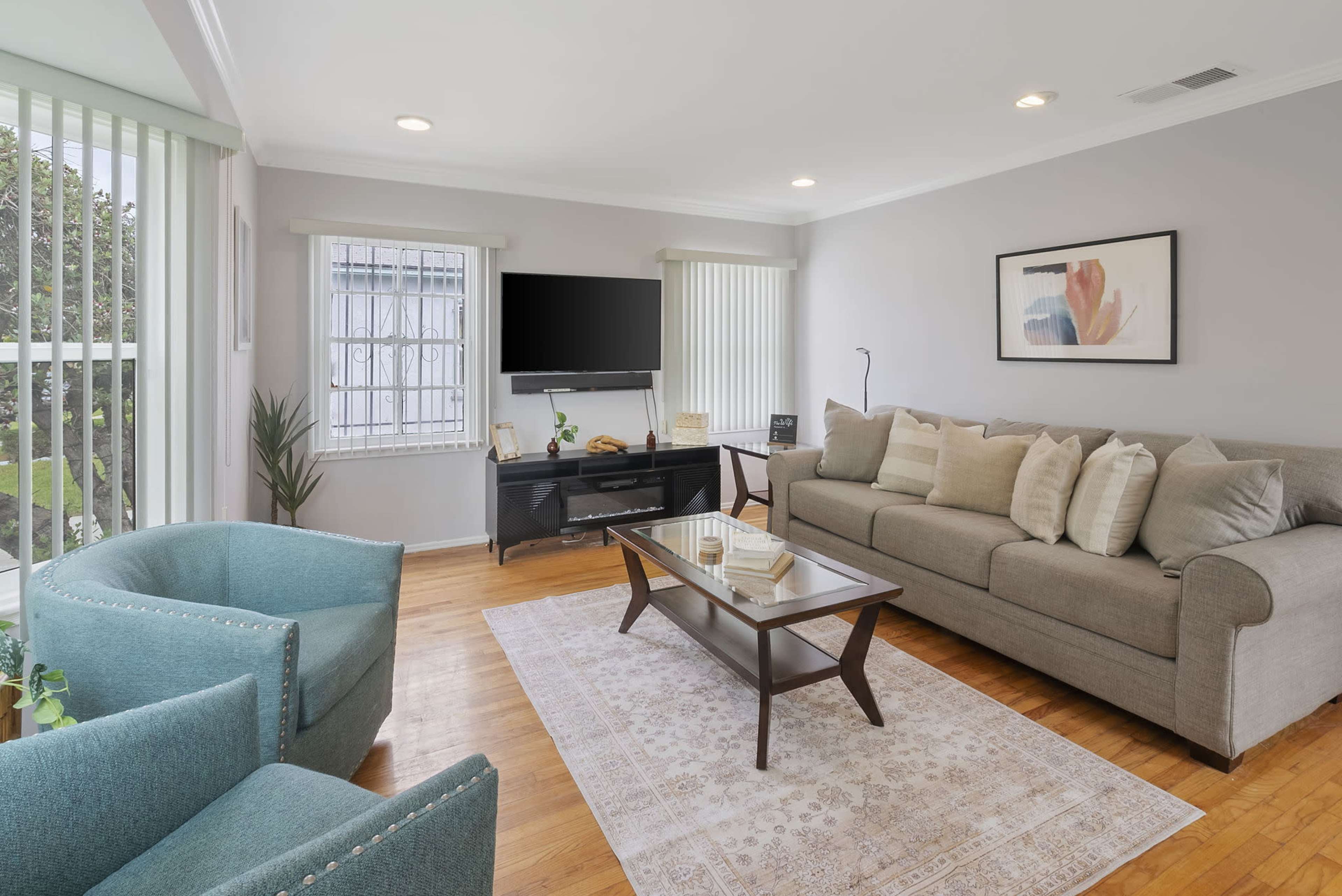 The image shows a living room featuring a sofa, two armchairs, a coffee table, a television mounted on the wall, and large windows with blinds.