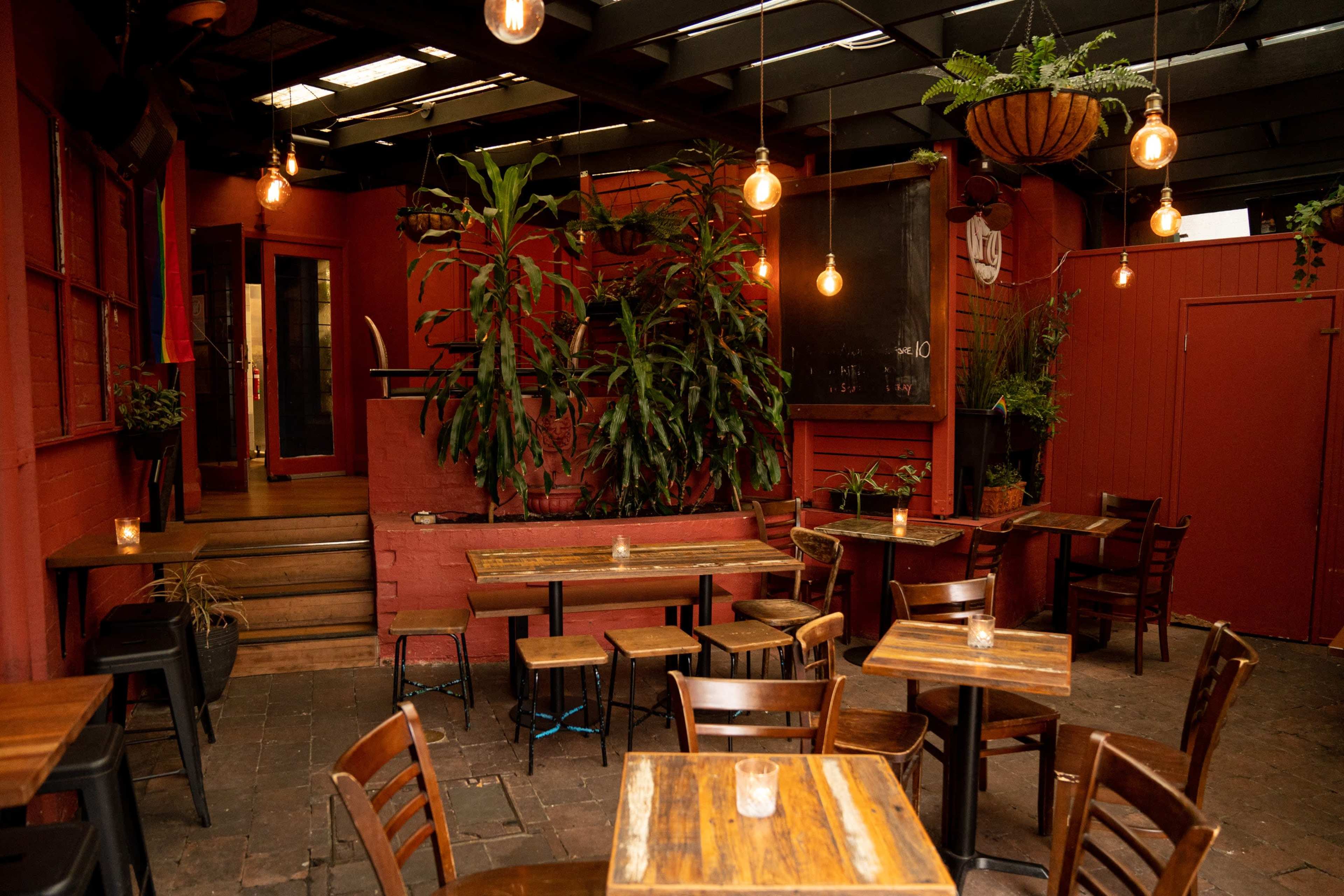 The image shows a cozy restaurant interior with wooden tables and chairs, illuminated by hanging lights and adorned with plants.