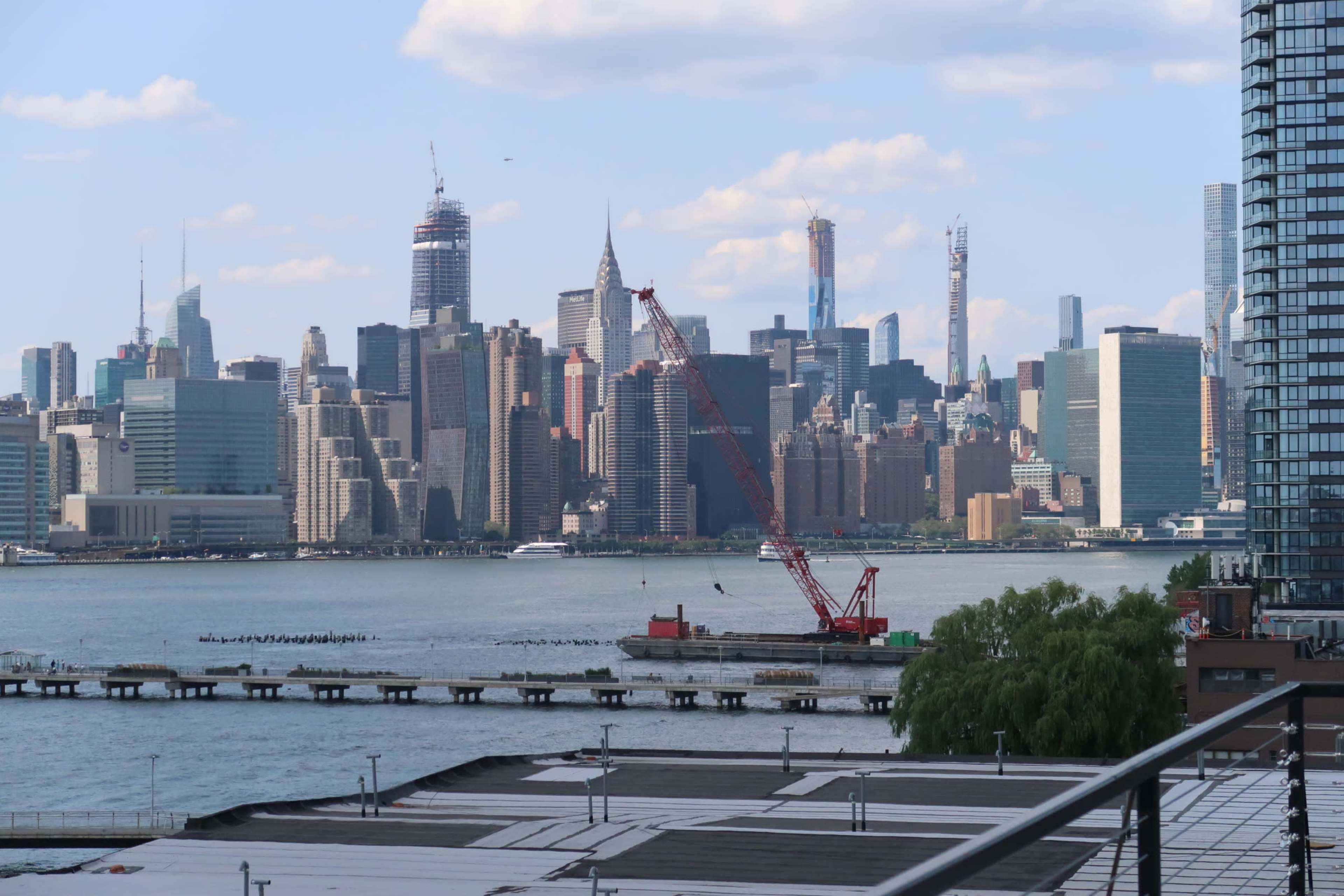 A view of the Manhattan skyline across the water, featuring skyscrapers and a red crane on a barge.