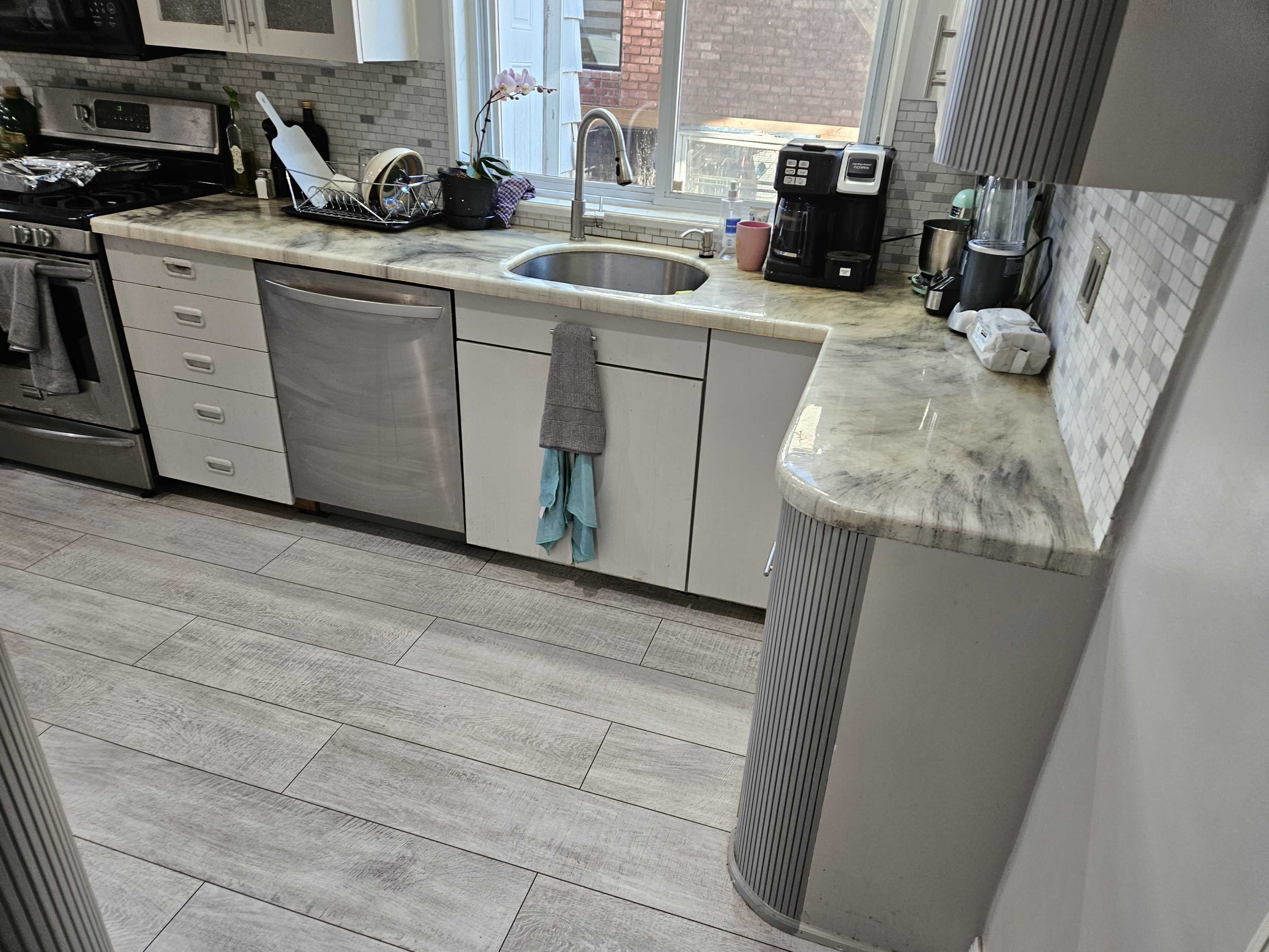 A modern kitchen with stainless steel appliances, a sink, and light-colored cabinetry arranged around a marble countertop.