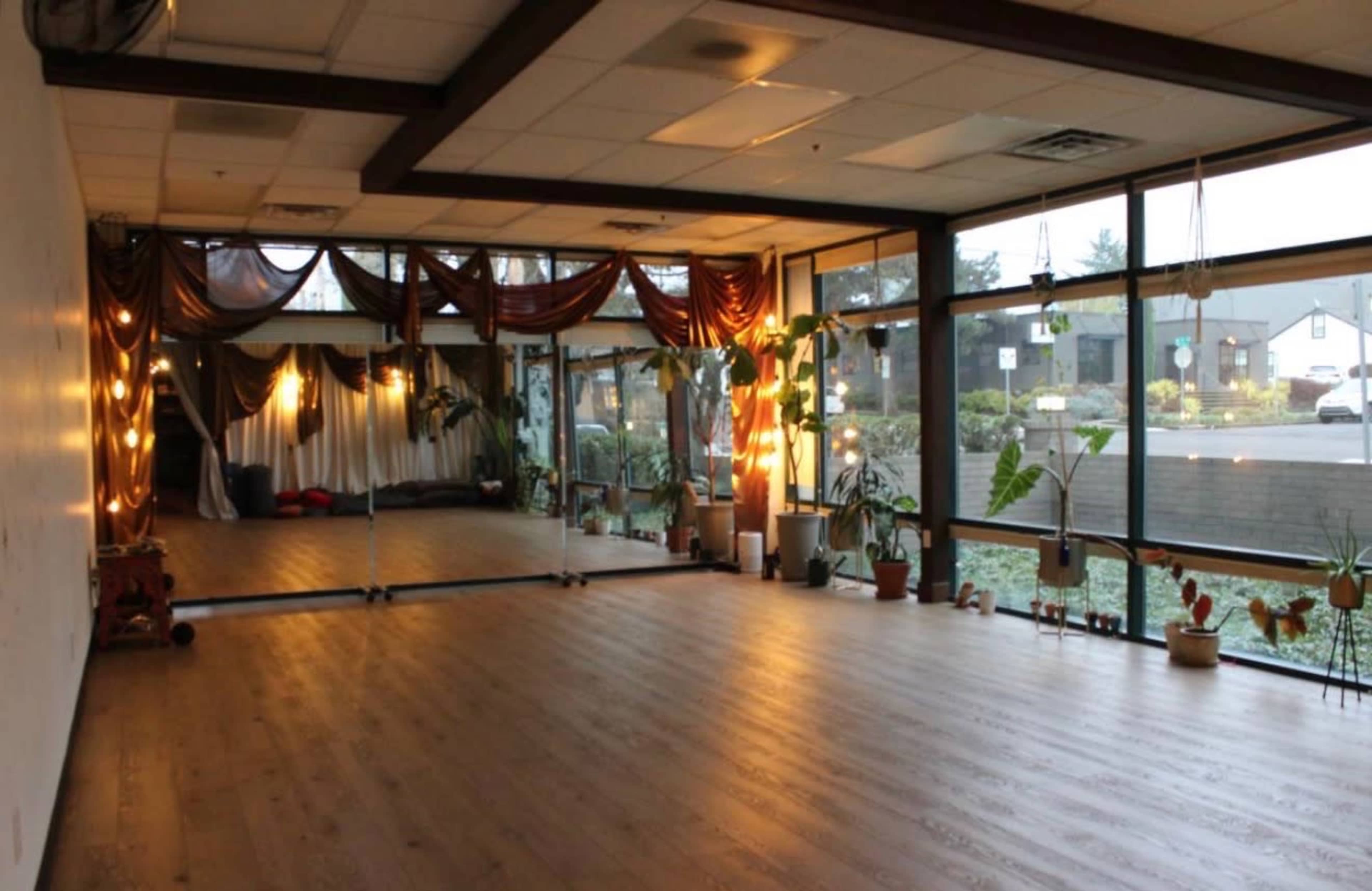 The image shows a spacious indoor area with large windows, wood flooring, plants, and decorative drapes, likely intended for yoga or dance classes.