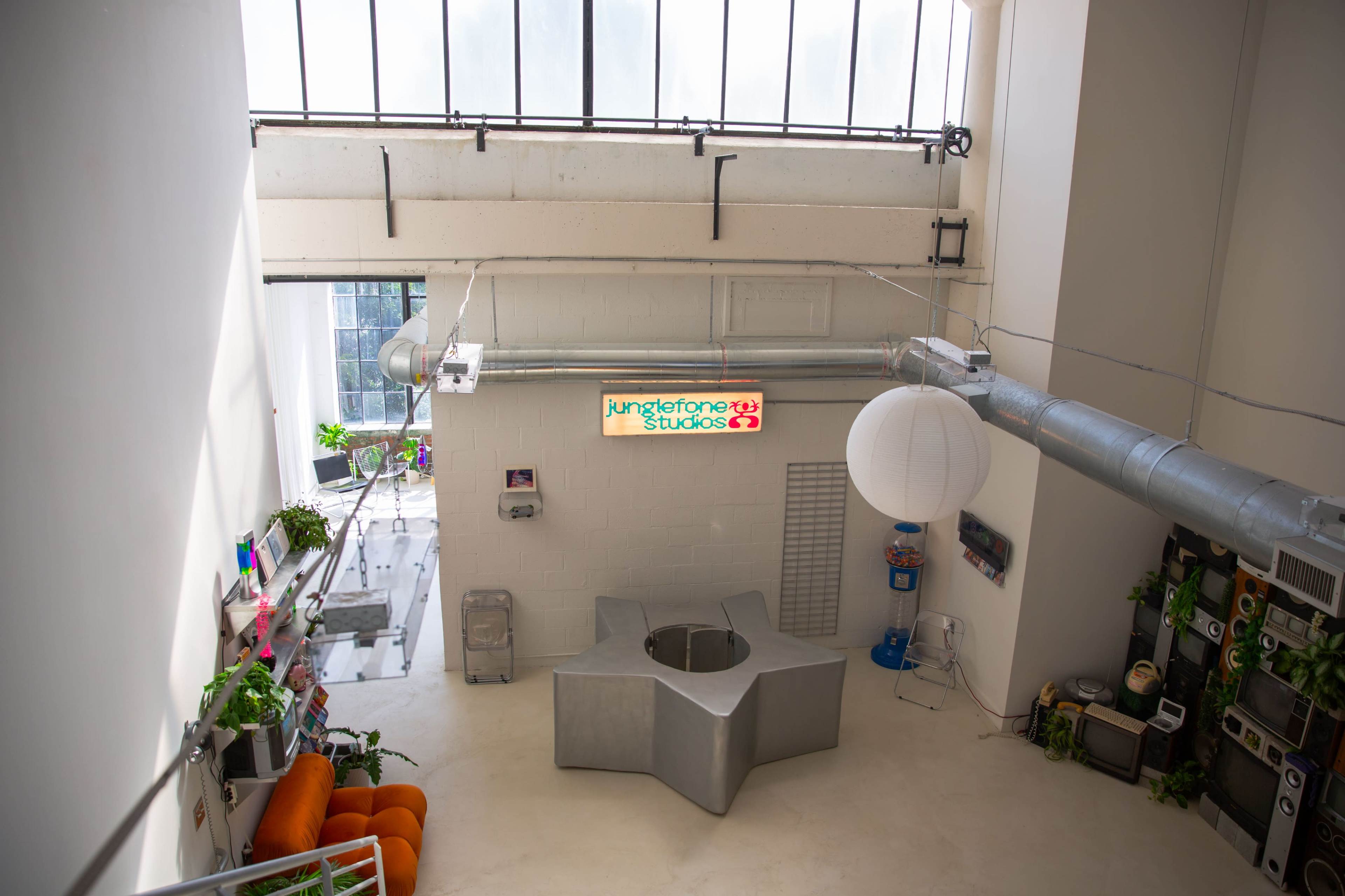 The image shows a spacious studio interior with large windows, a circular seating area, and various plants along the walls.