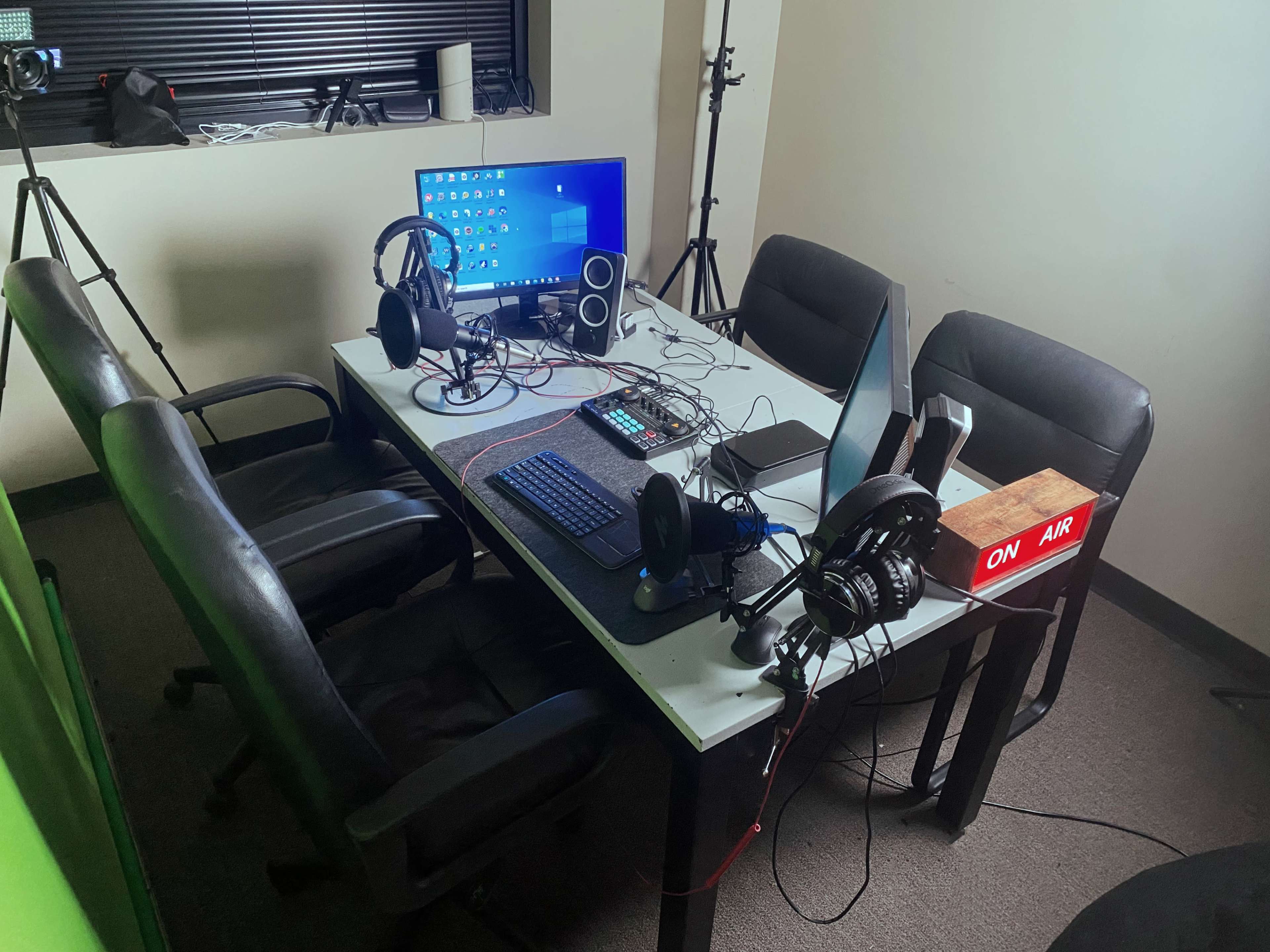A media production setup with a table, four chairs, a computer monitor, audio equipment, and a "ON AIR" sign.
