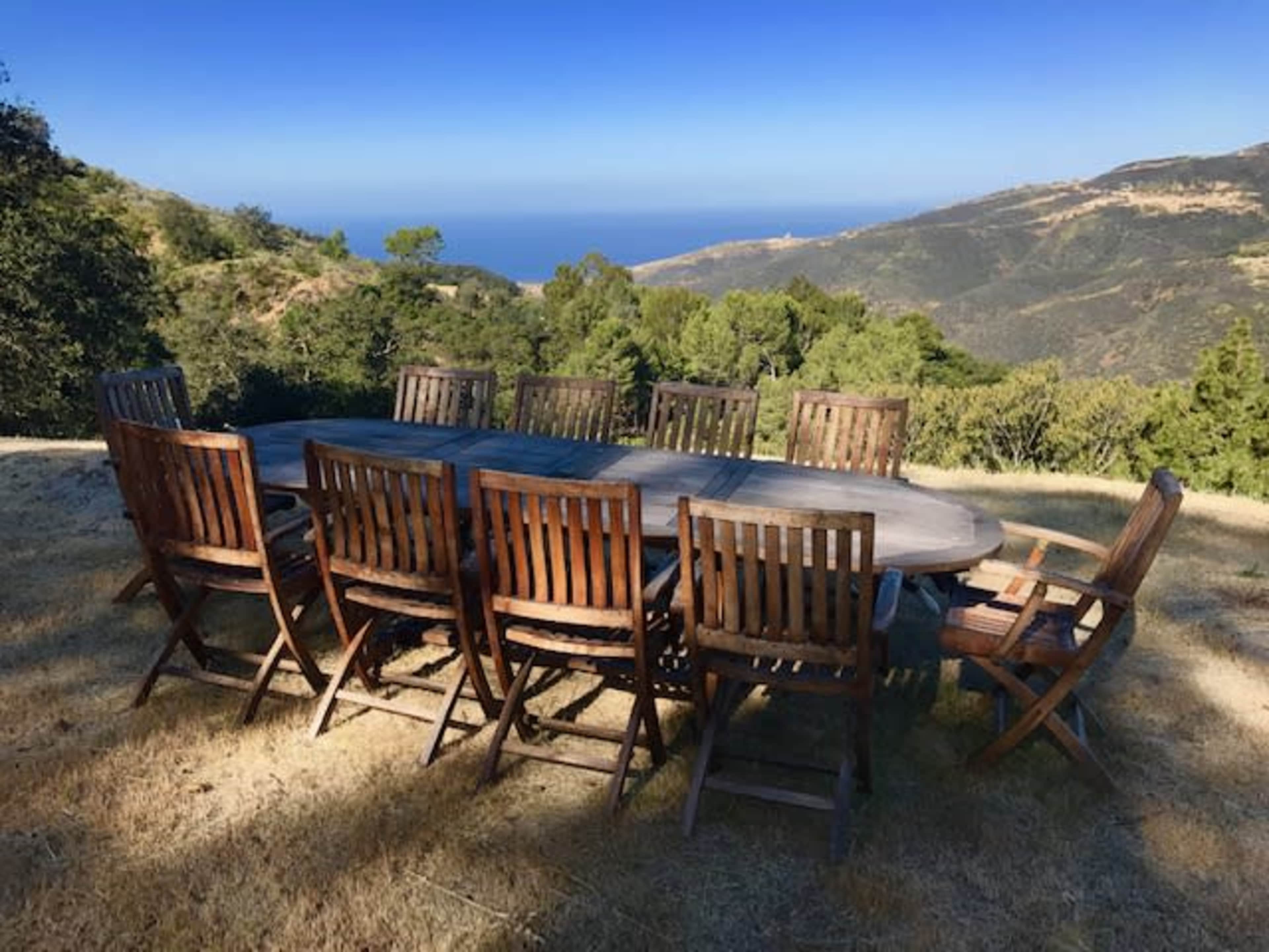 A long wooden dining table is surrounded by wooden chairs, set on a grassy area overlooking a mountainous landscape and ocean in the distance.