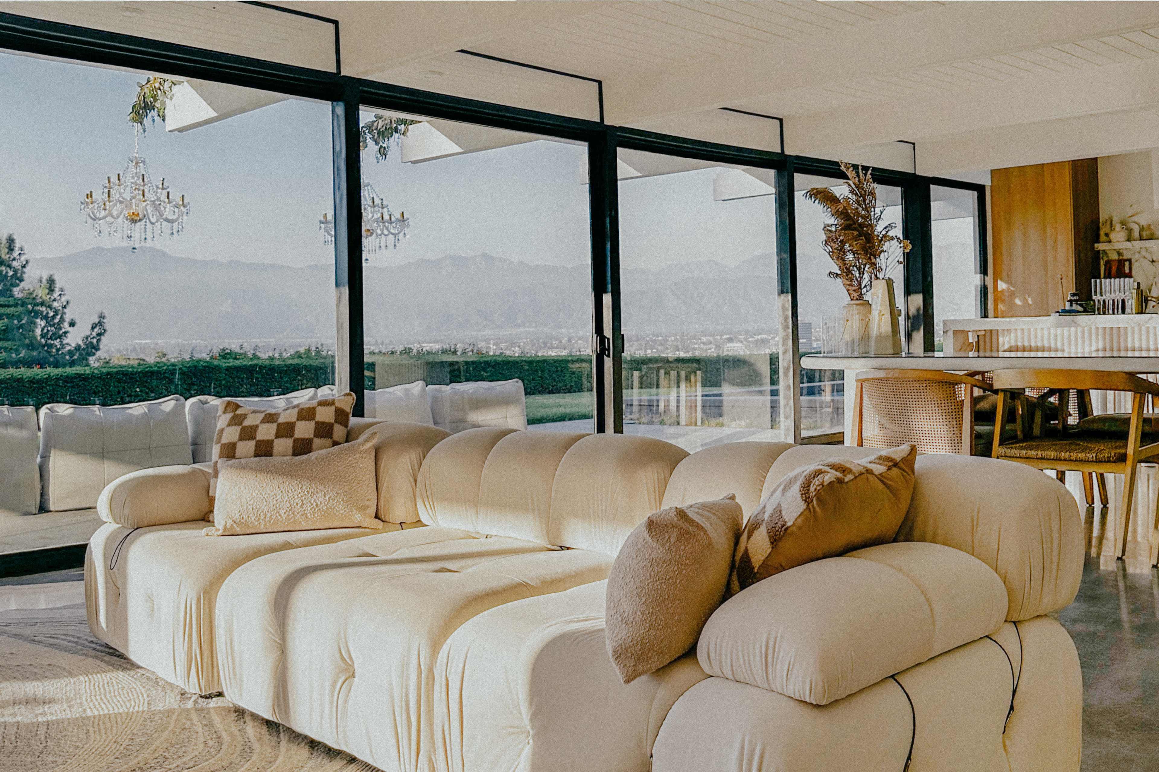 A spacious living area features a white, plush sofa with decorative pillows, large glass windows showcasing a mountain view, and a modern dining space nearby.