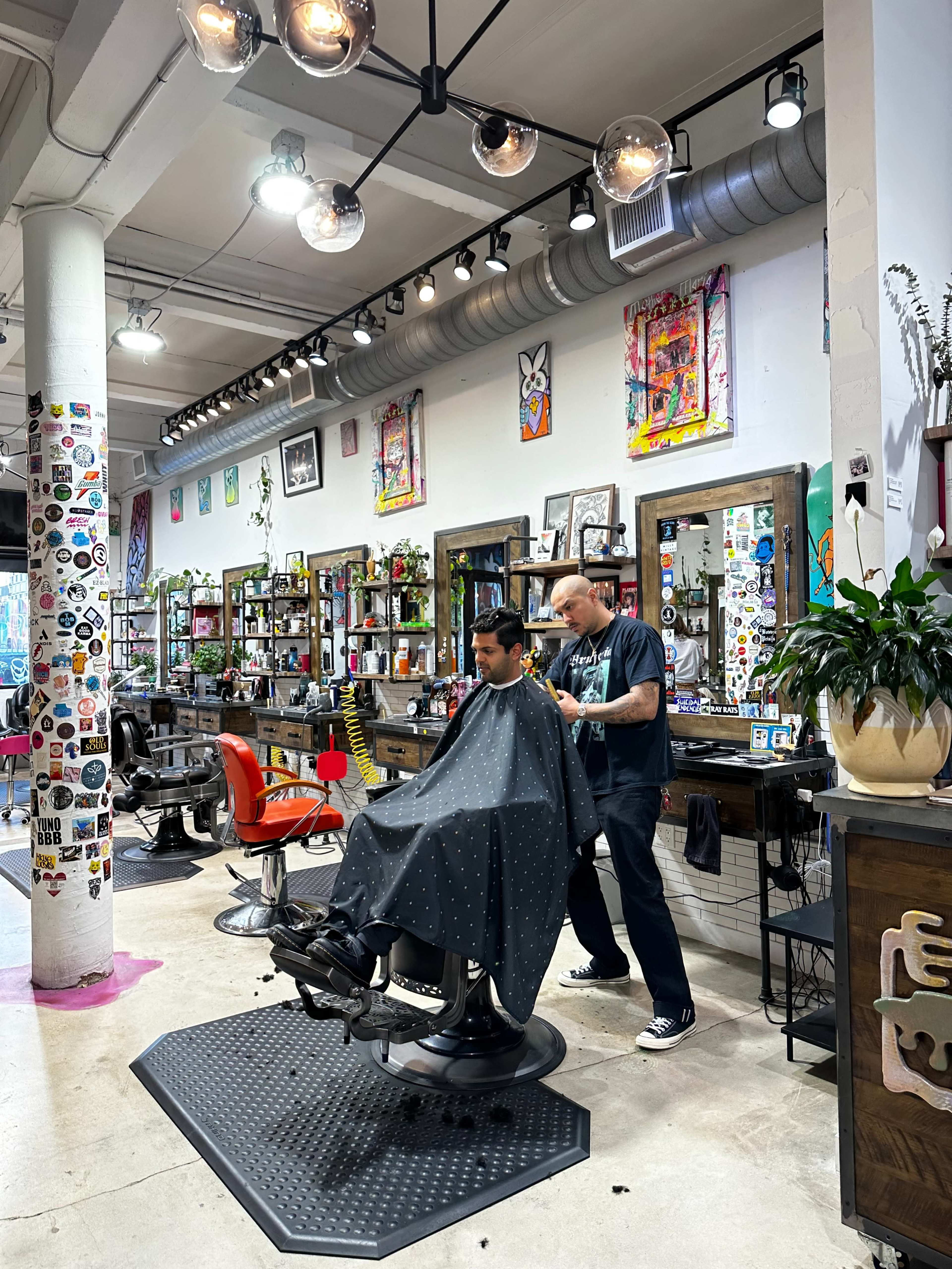 A barber is cutting a customer's hair in a modern, decorated barbershop with colorful artwork on the walls.
