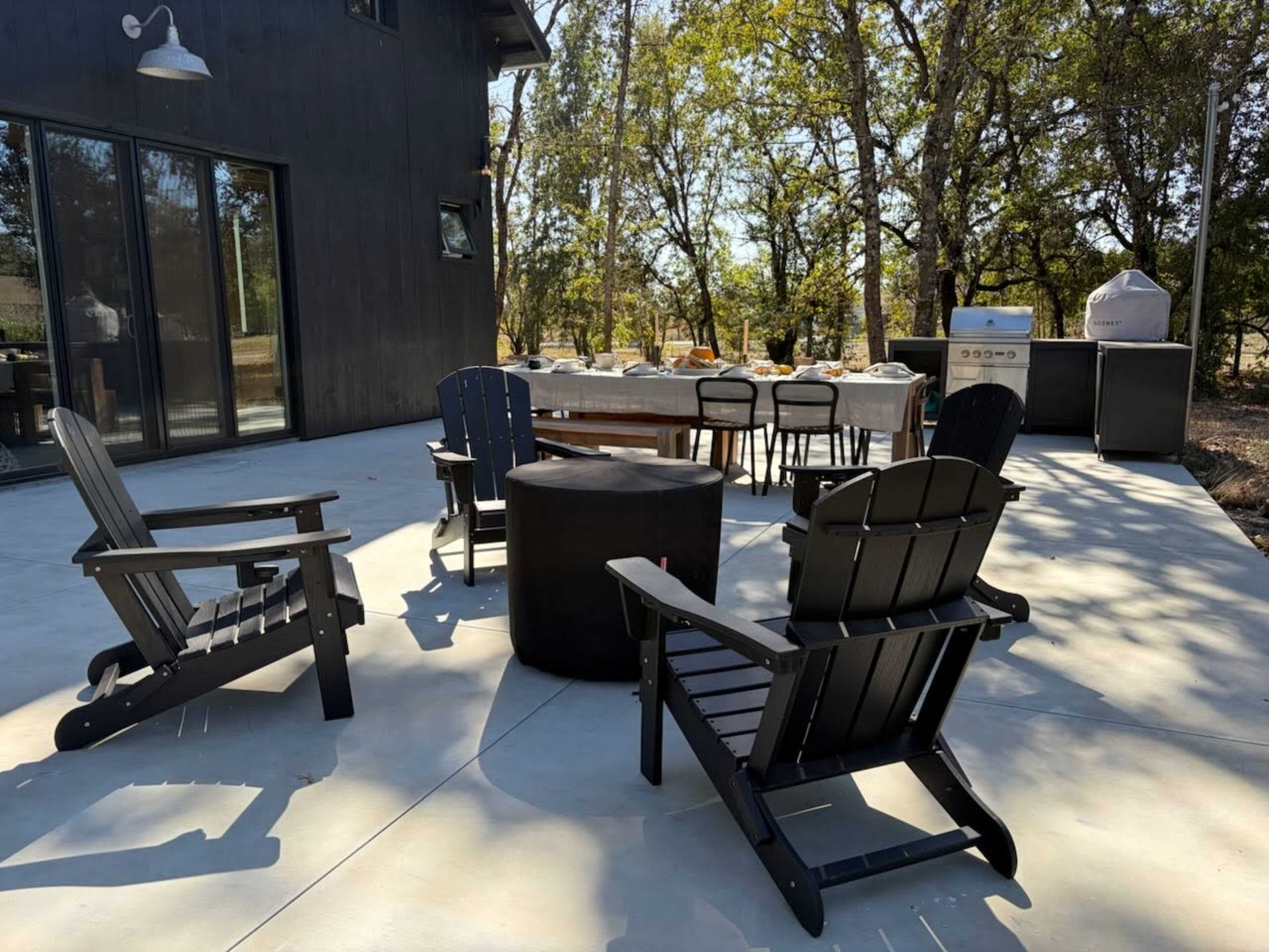 A patio area features black wooden Adirondack chairs arranged around a circular table, with a dining table set in the background under trees.