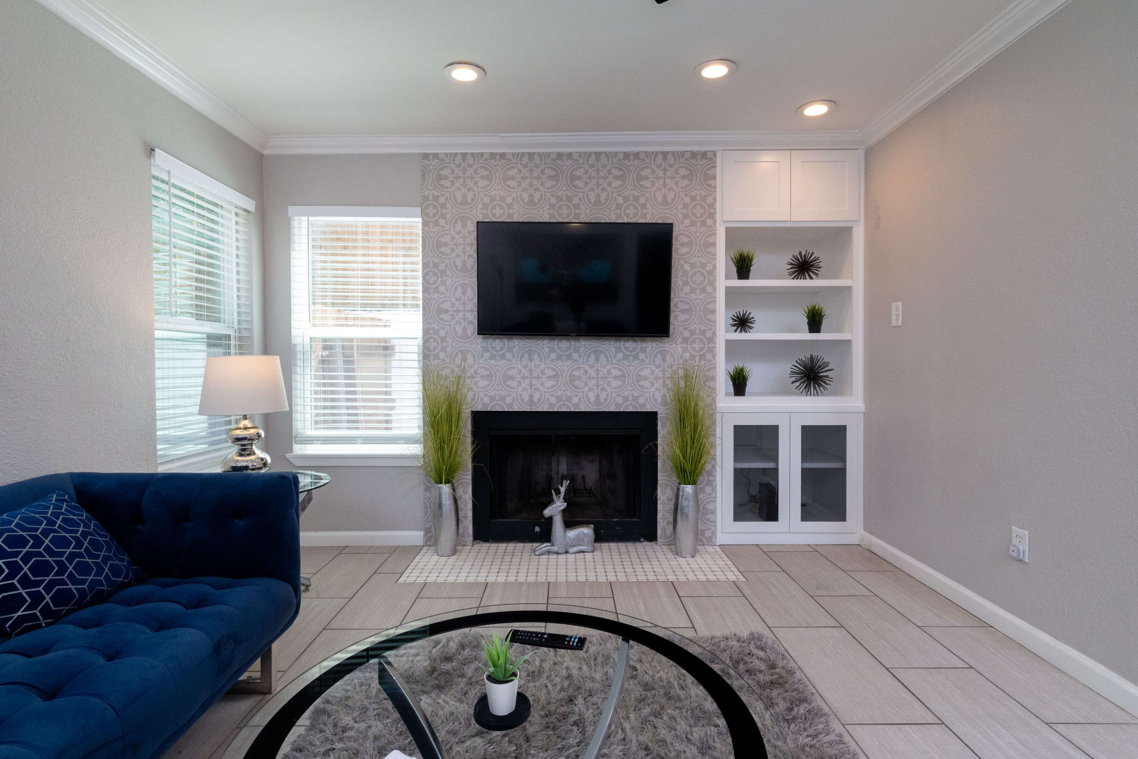 The image shows a living room with a blue sofa, a mounted television above a fireplace, and a white shelving unit displaying decorative items.