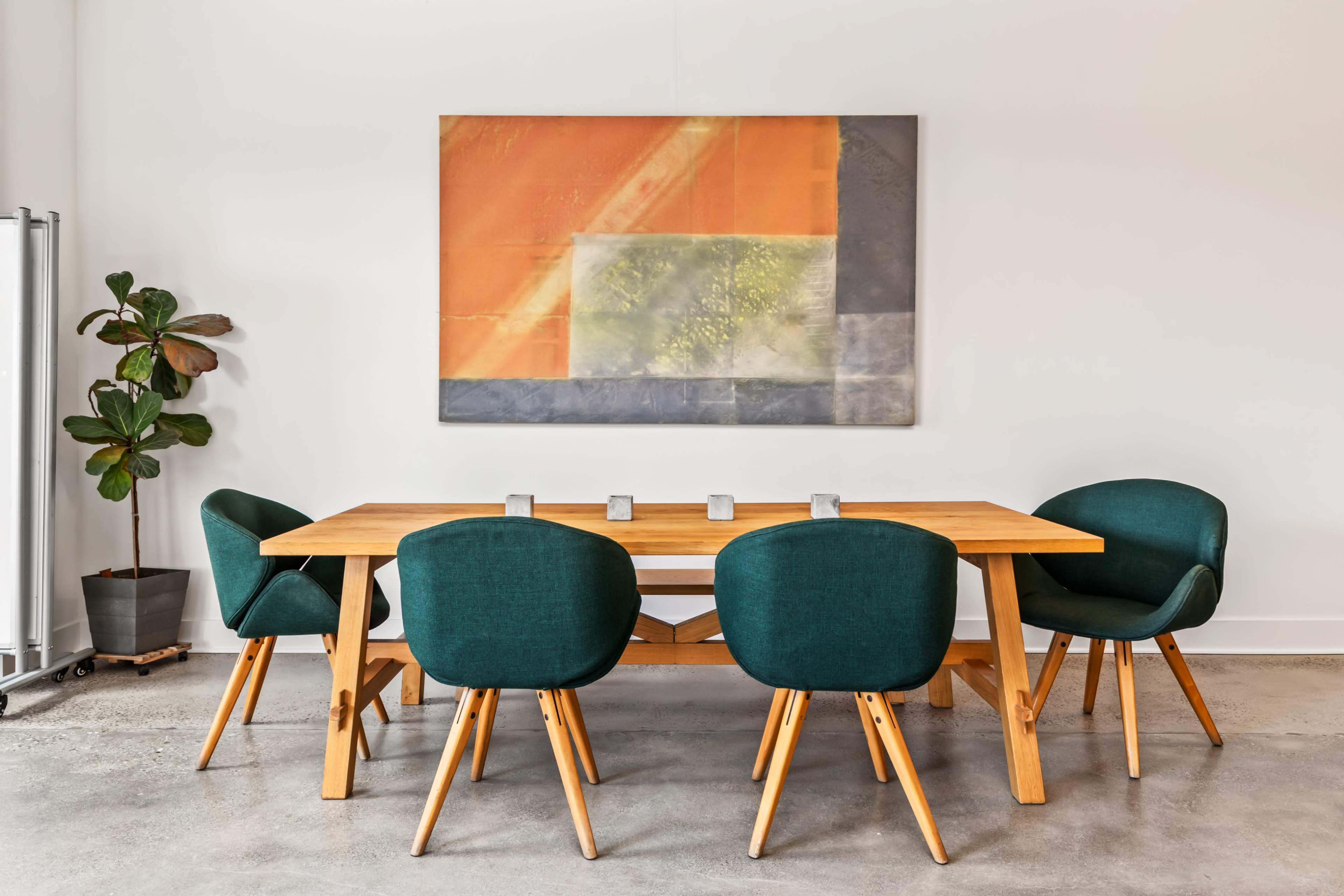 A wooden dining table with four green chairs is set against a white wall featuring a large abstract painting and a potted plant.