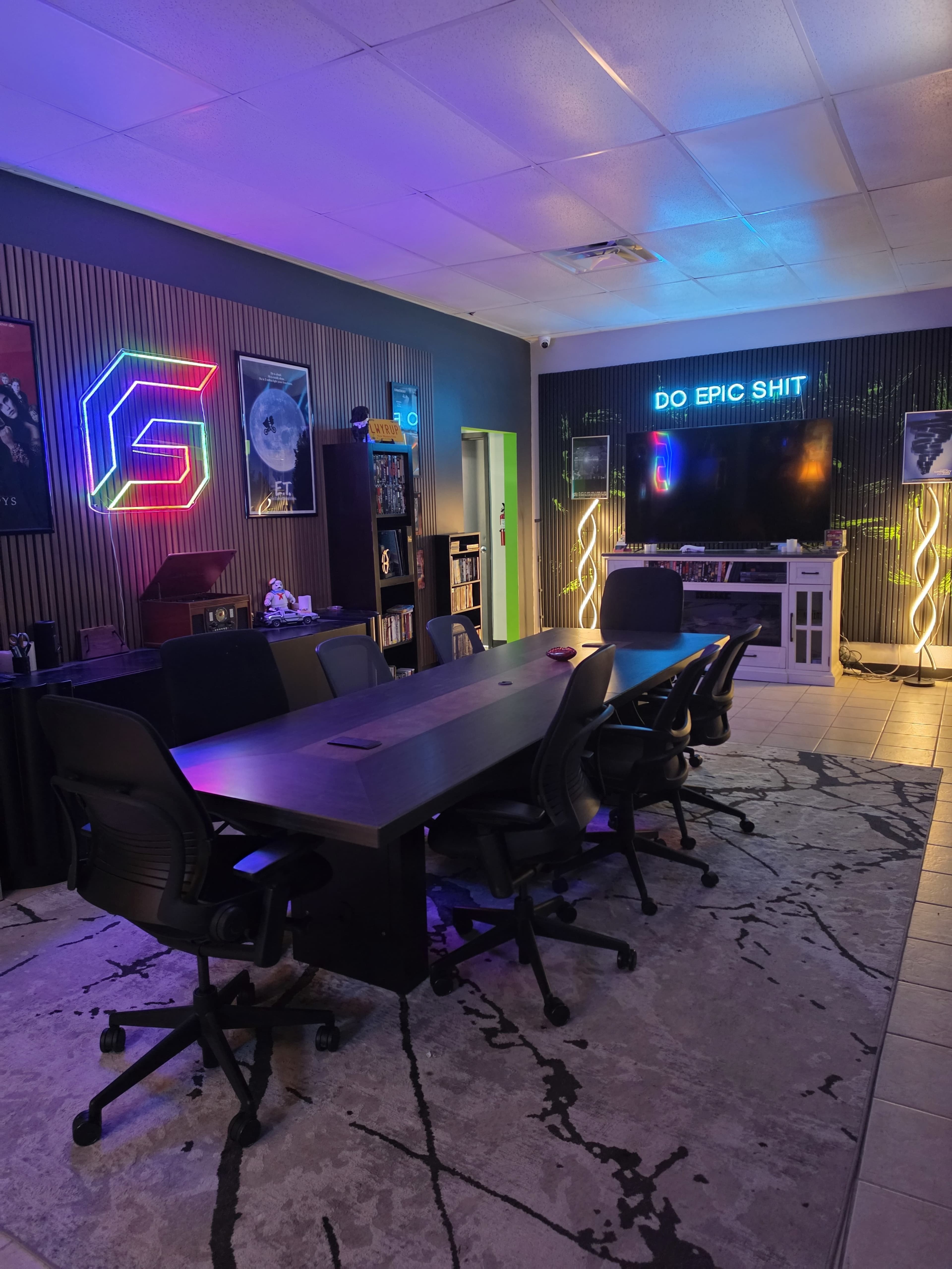 The image shows a modern conference room featuring a large table, several ergonomic chairs, colorful LED lighting, and a large screen displaying the phrase "DO EPIC SHIT."
