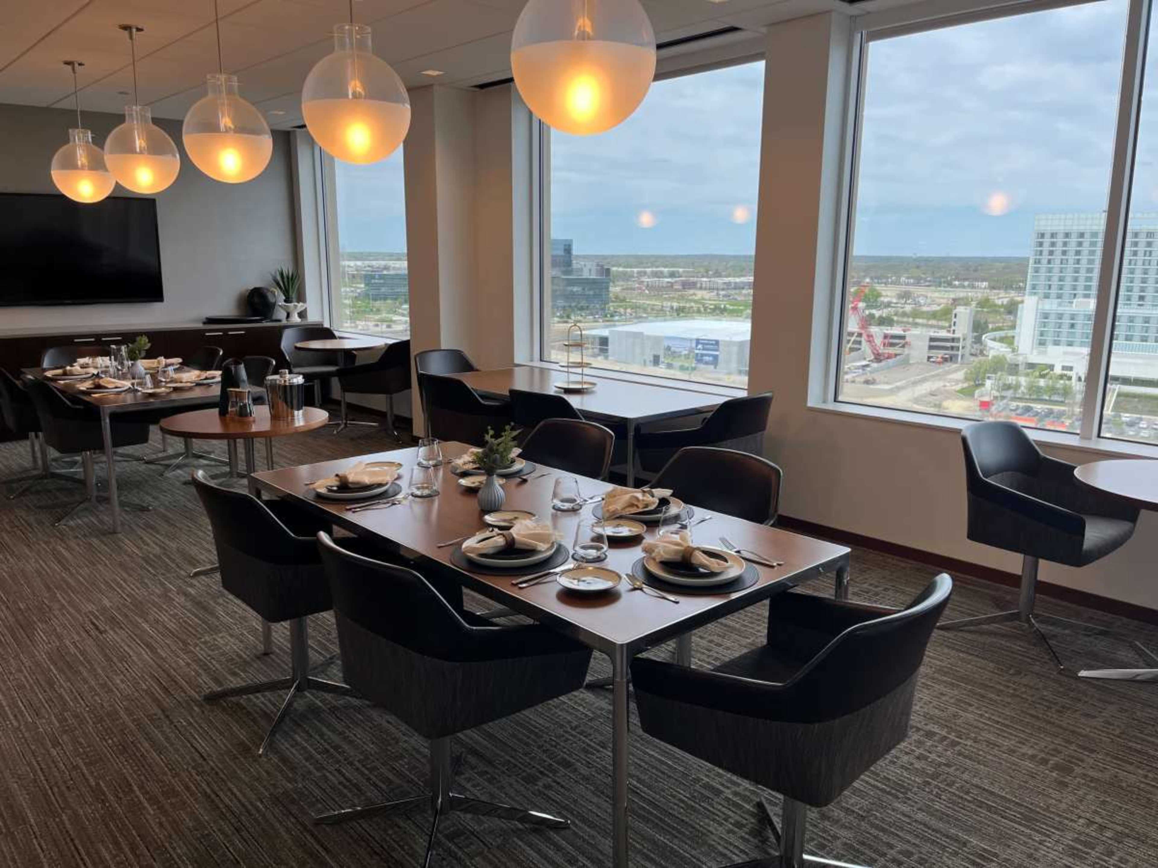 A modern dining area features elegantly set tables with chairs, large windows offering a view of the city, and pendant lighting hanging from the ceiling.